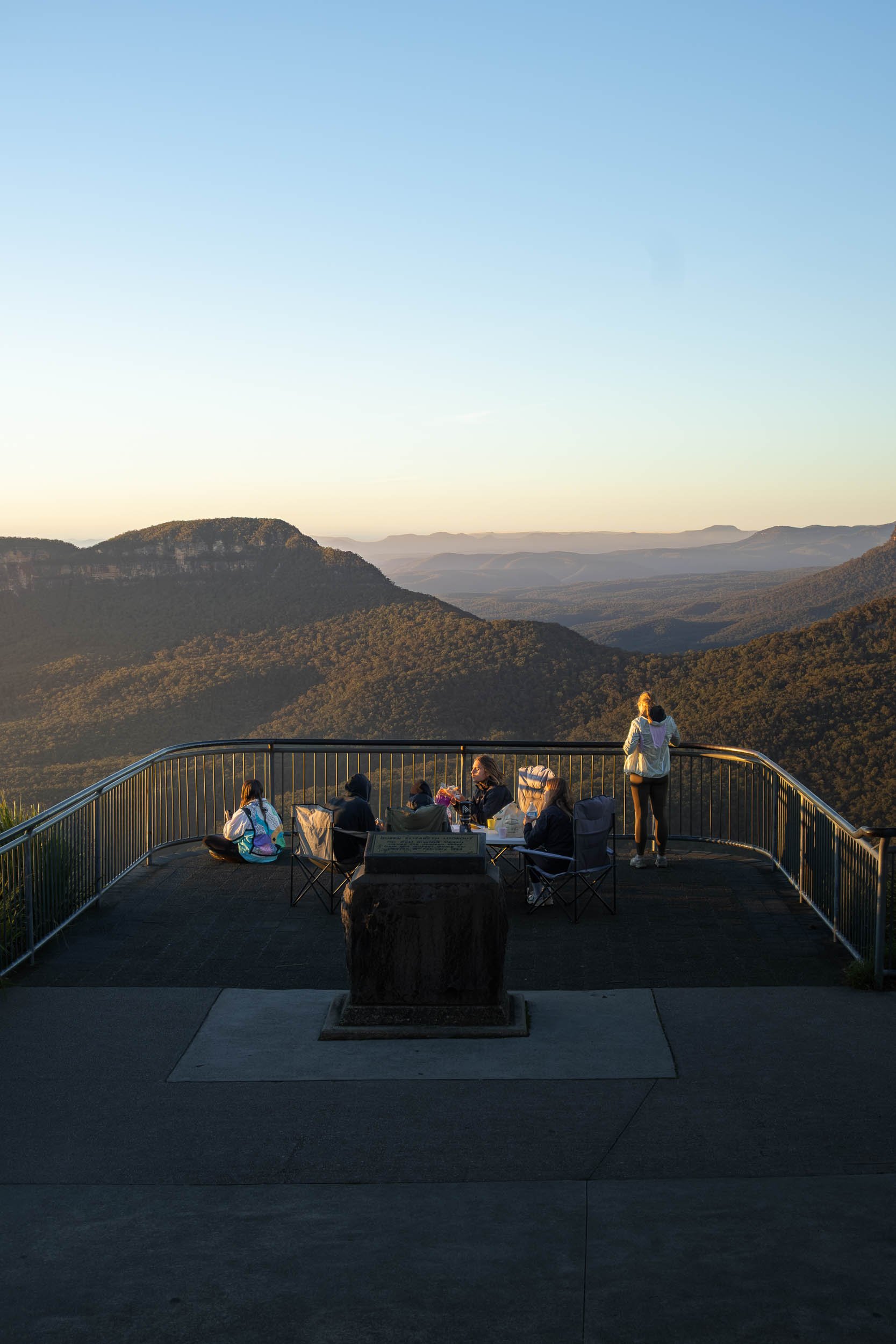 Group of people sitting and standing on an observation deck overlooking a mountain landscape at sunset.