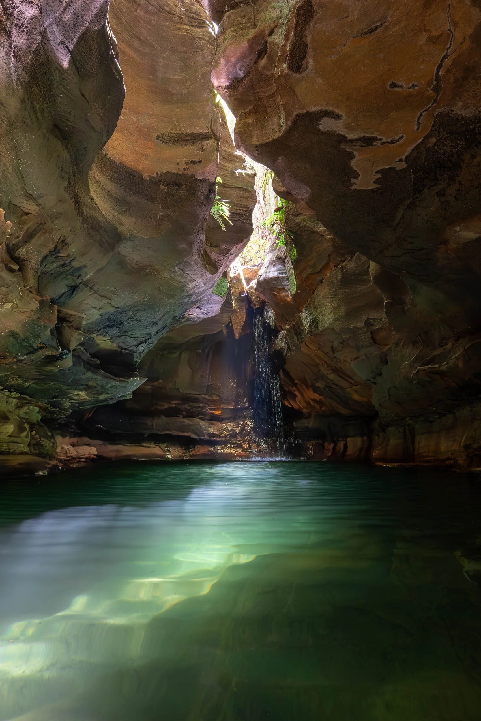 A narrow canyon with smooth, multi-colored rock walls and a small waterfall flowing into a clear green pool of water.