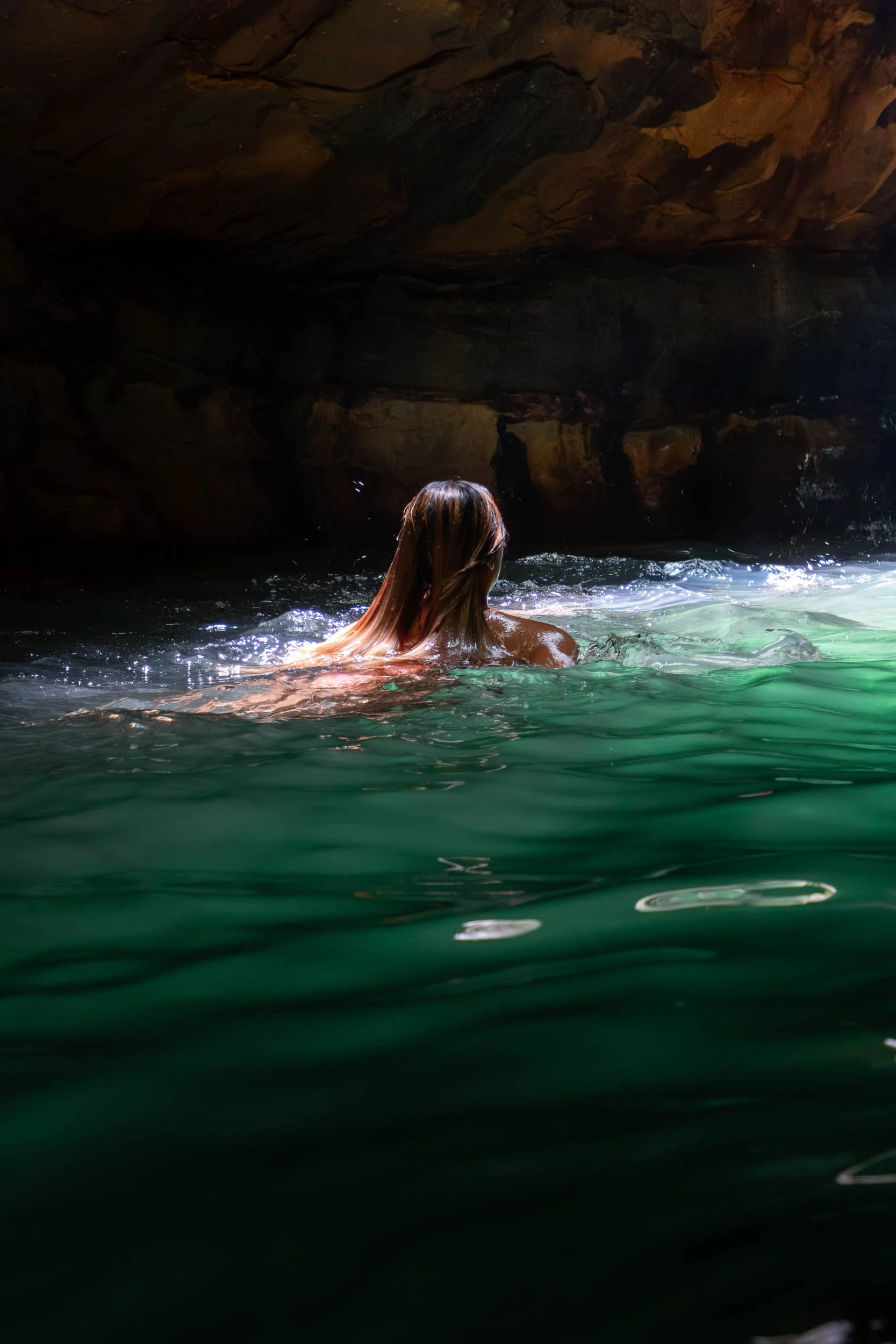 A person with long, wet hair swimming in a dark cave or tunnel with greenish water and rocky walls.