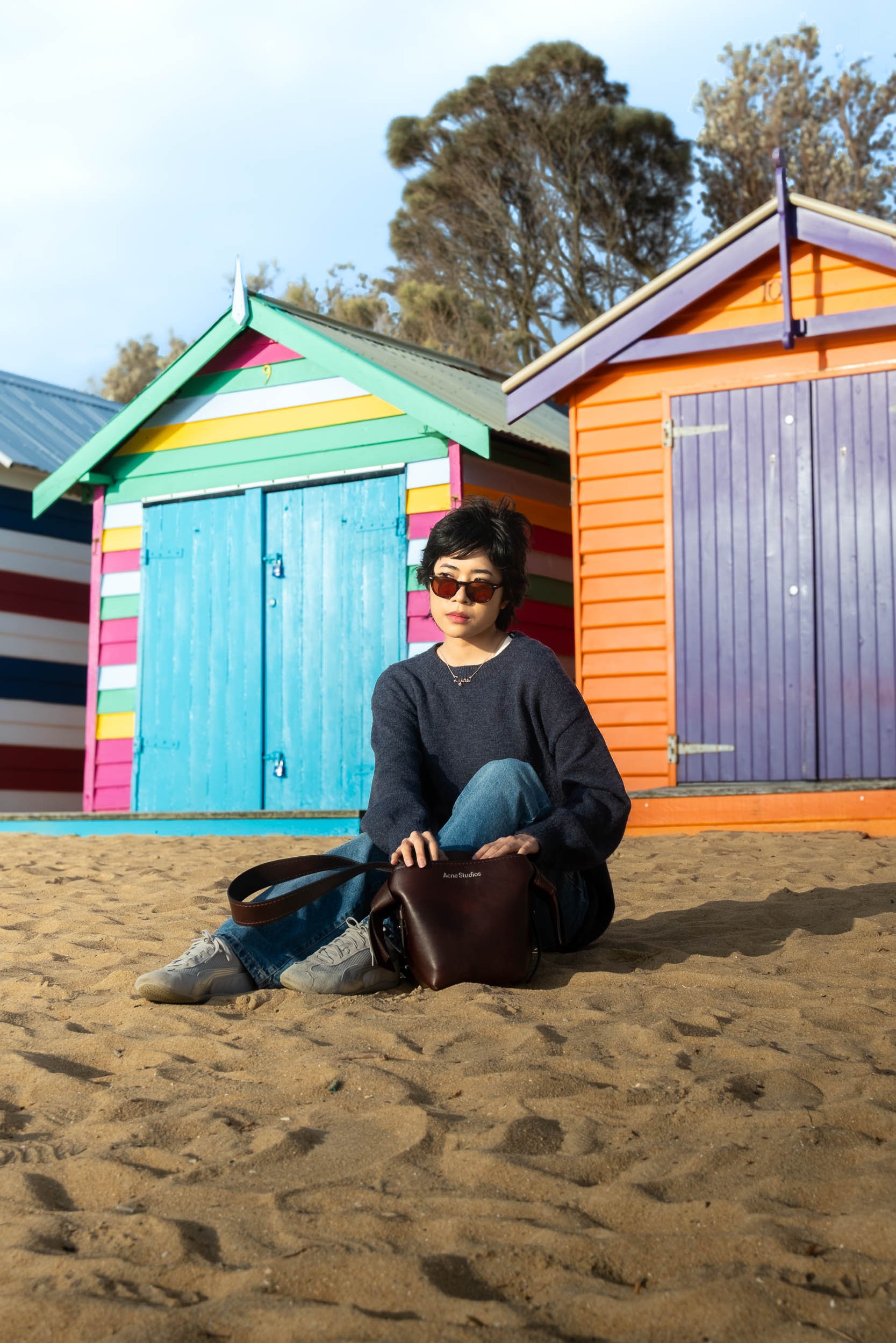 A woman sitting on sandy ground with colorful beach huts behind her, wearing sunglasses, a dark sweater, jeans, and sneakers, holding a brown bag.