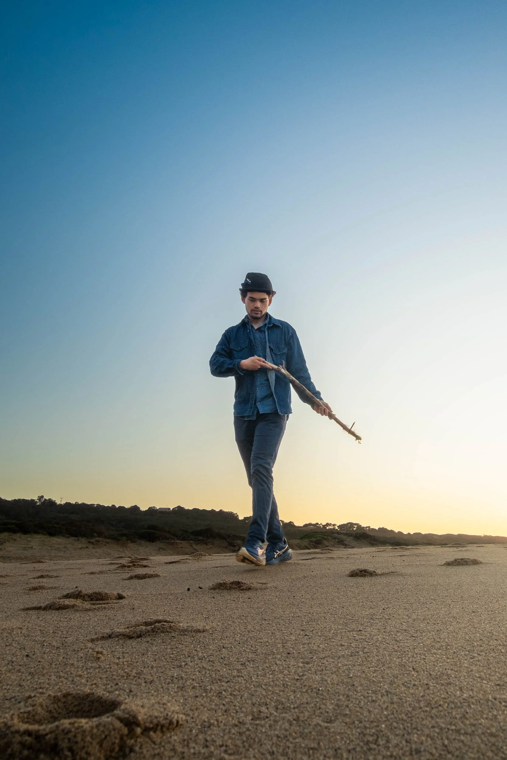 A man walking on a sandy beach during sunset, holding a stick and wearing a denim jacket, dark pants, sneakers, and a hat.