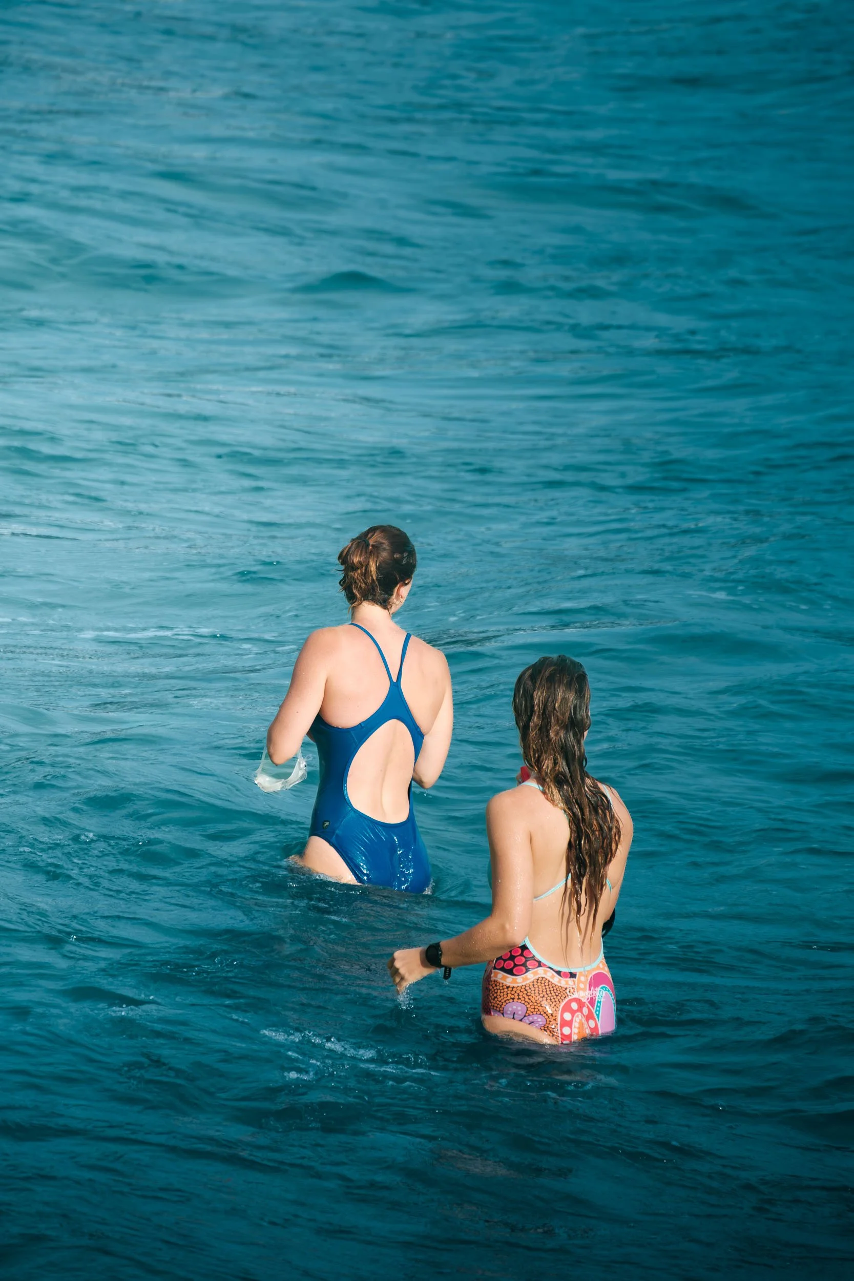 Two women are standing in the ocean, with one facing away and the other slightly turned. They are wearing swimsuits and are surrounded by blue water, with gentle waves.