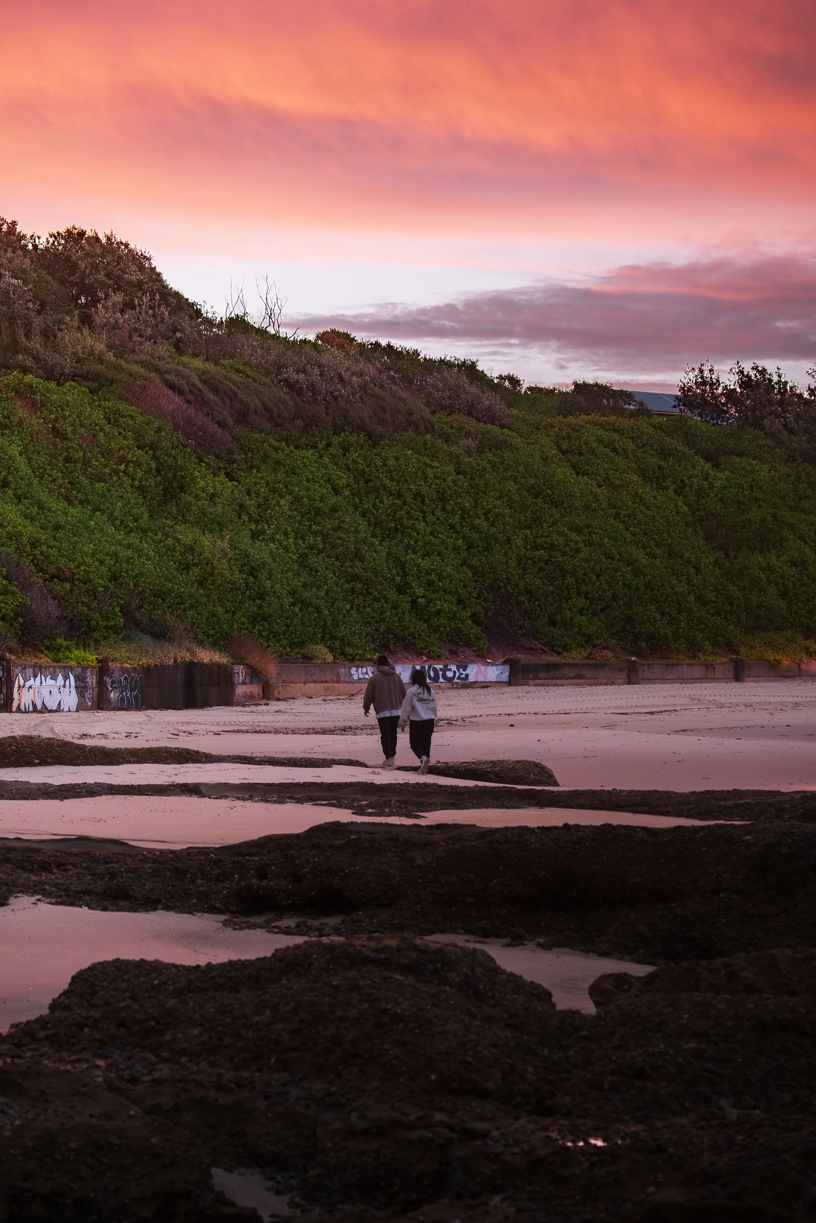 Two people walking along a beach at sunset with a colorful sky and green hillside in the background.