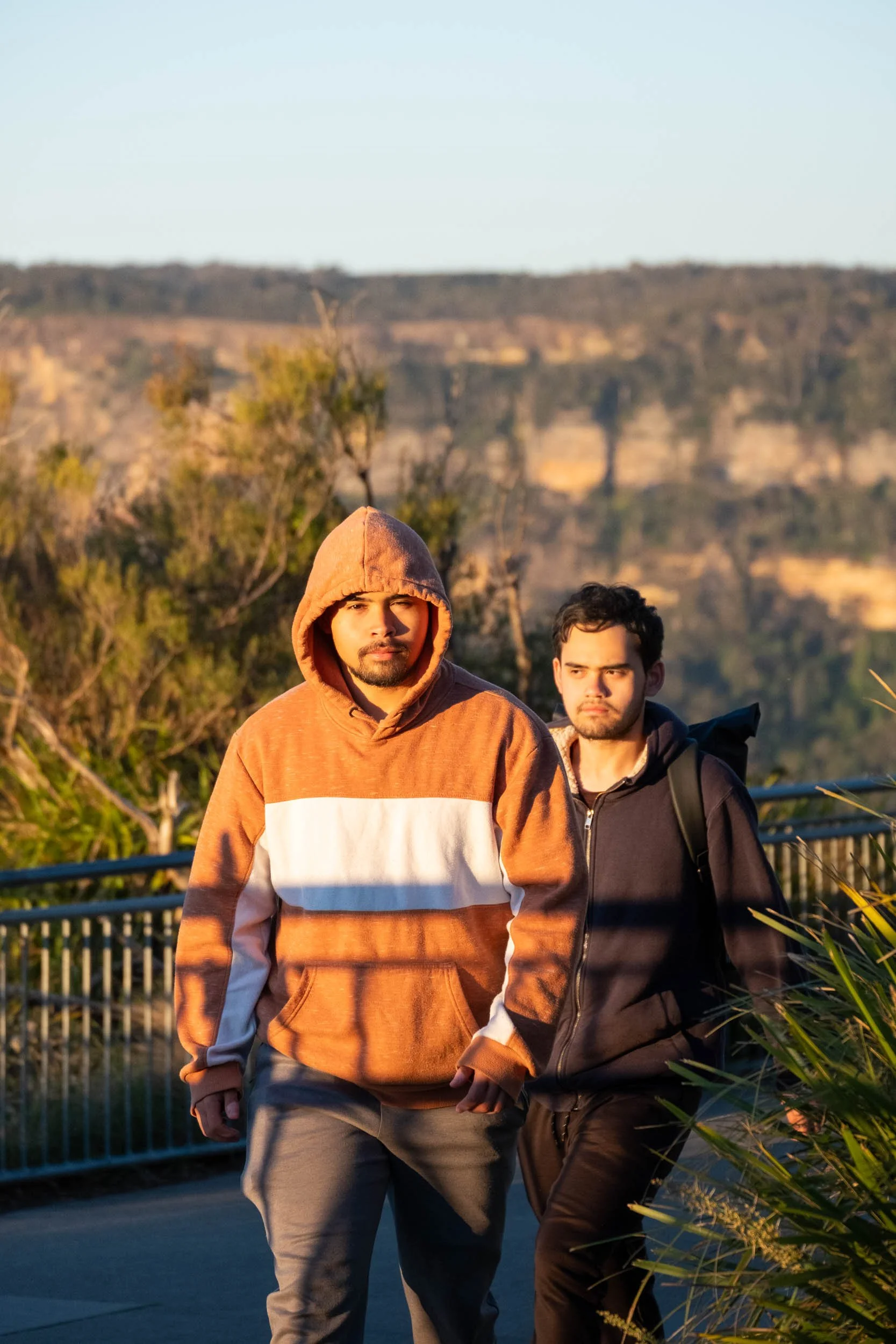 Two young men walking outdoors during sunset, with trees and cliffs in the background. One is wearing a brown hoodie with a white stripe, and the other is wearing a dark jacket with a backpack.