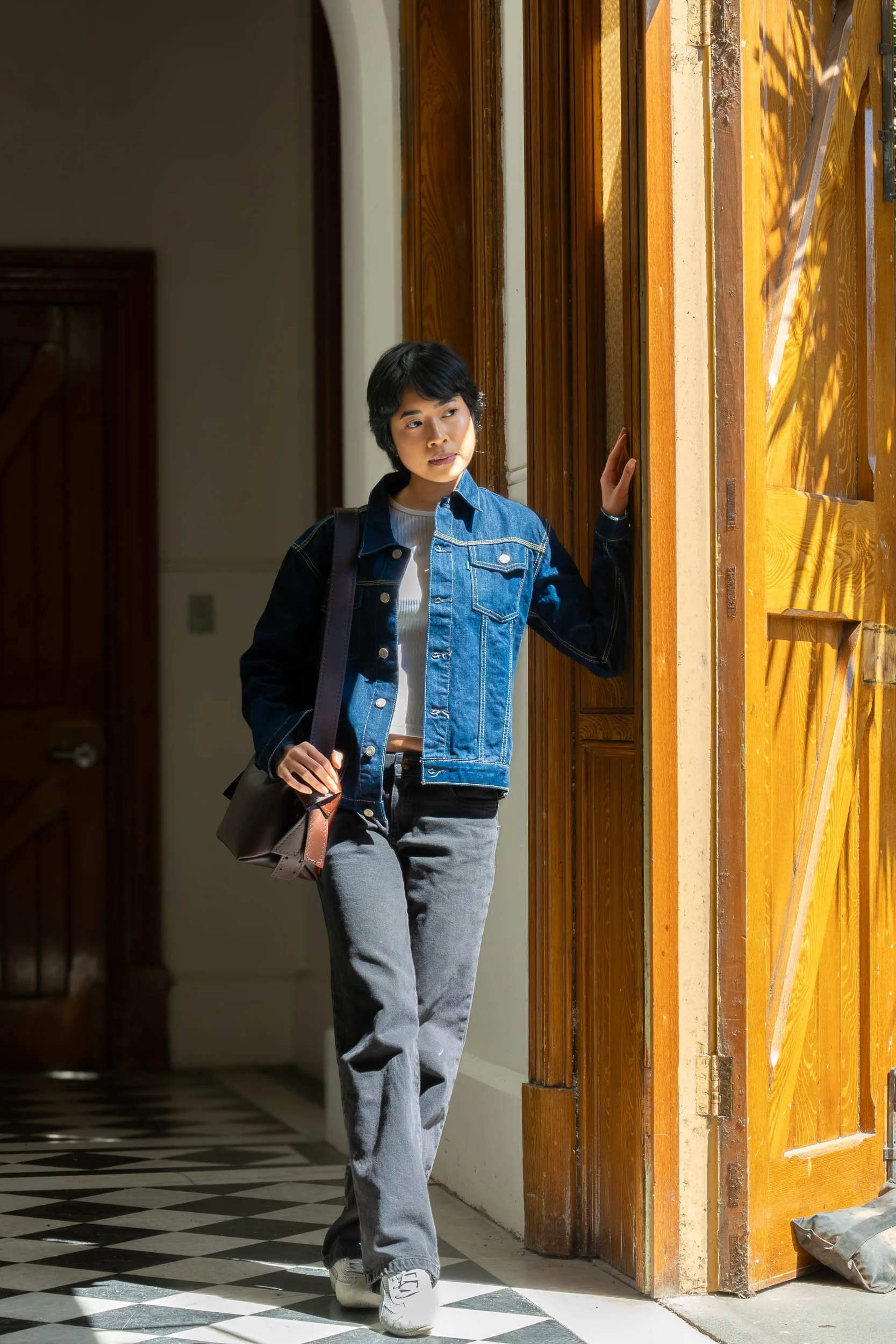 A young woman with short black hair, wearing a denim jacket, white shirt, gray jeans, and white sneakers, standing by a wooden door with sunlight streaming through, holding a brown shoulder bag.
