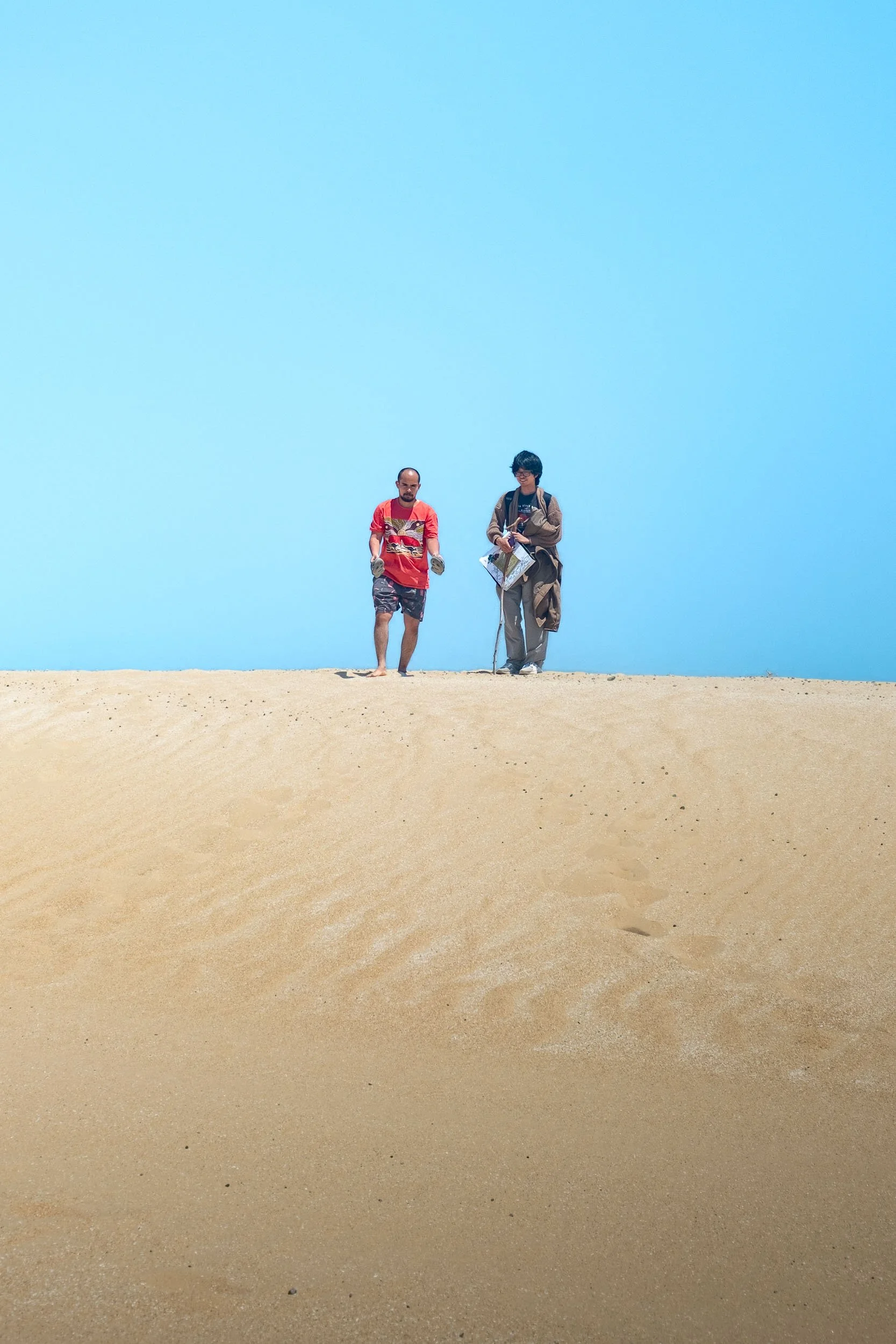 Two people walking on sand dunes against a clear blue sky, one carrying a backpack and the other holding a map.