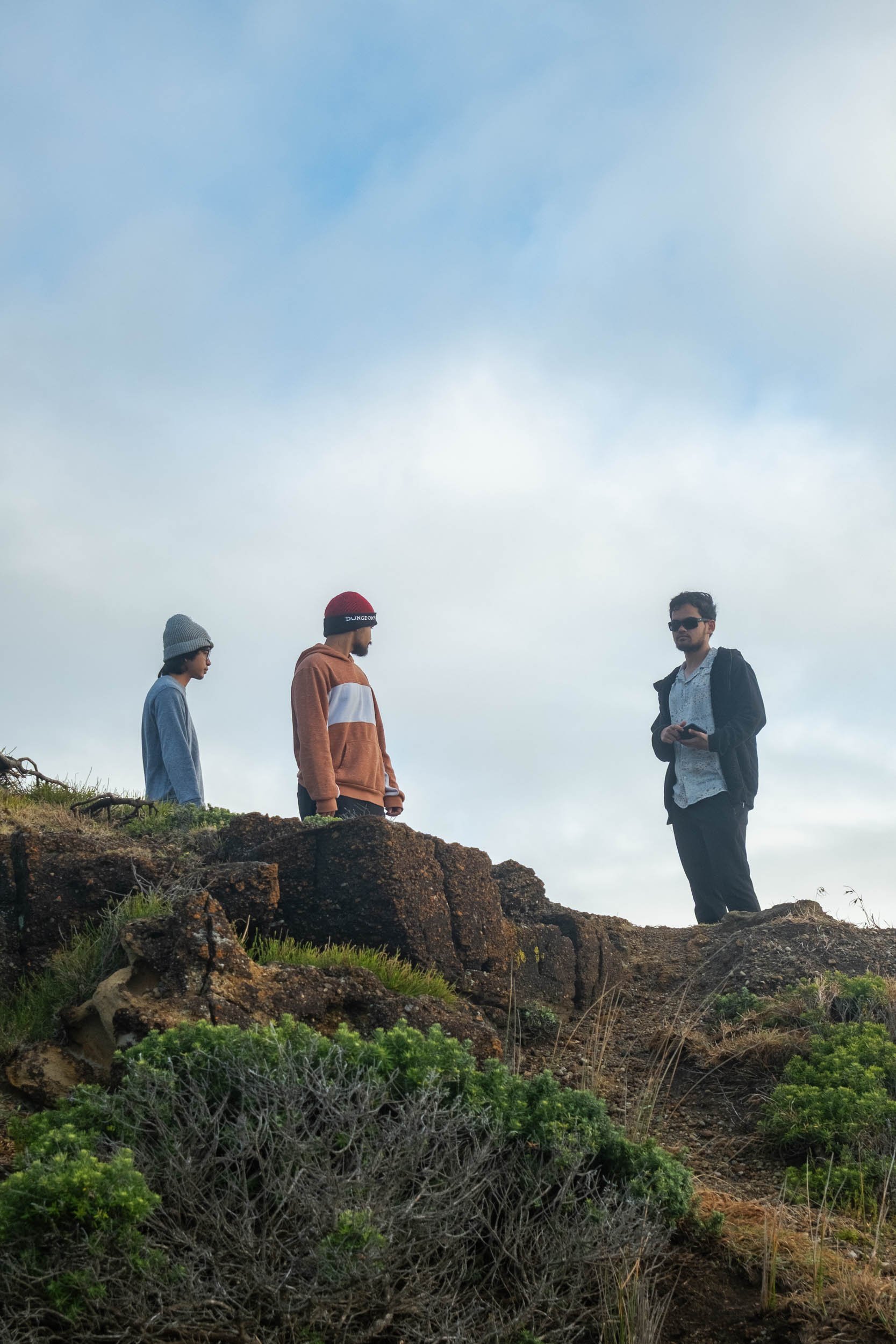 Three men standing on rocky terrain outdoors, with a cloudy sky in the background. One man is on the right holding a phone, wearing sunglasses and a dark jacket. The other two are on the left, one wearing a grey beanie and blue shirt, and the other i