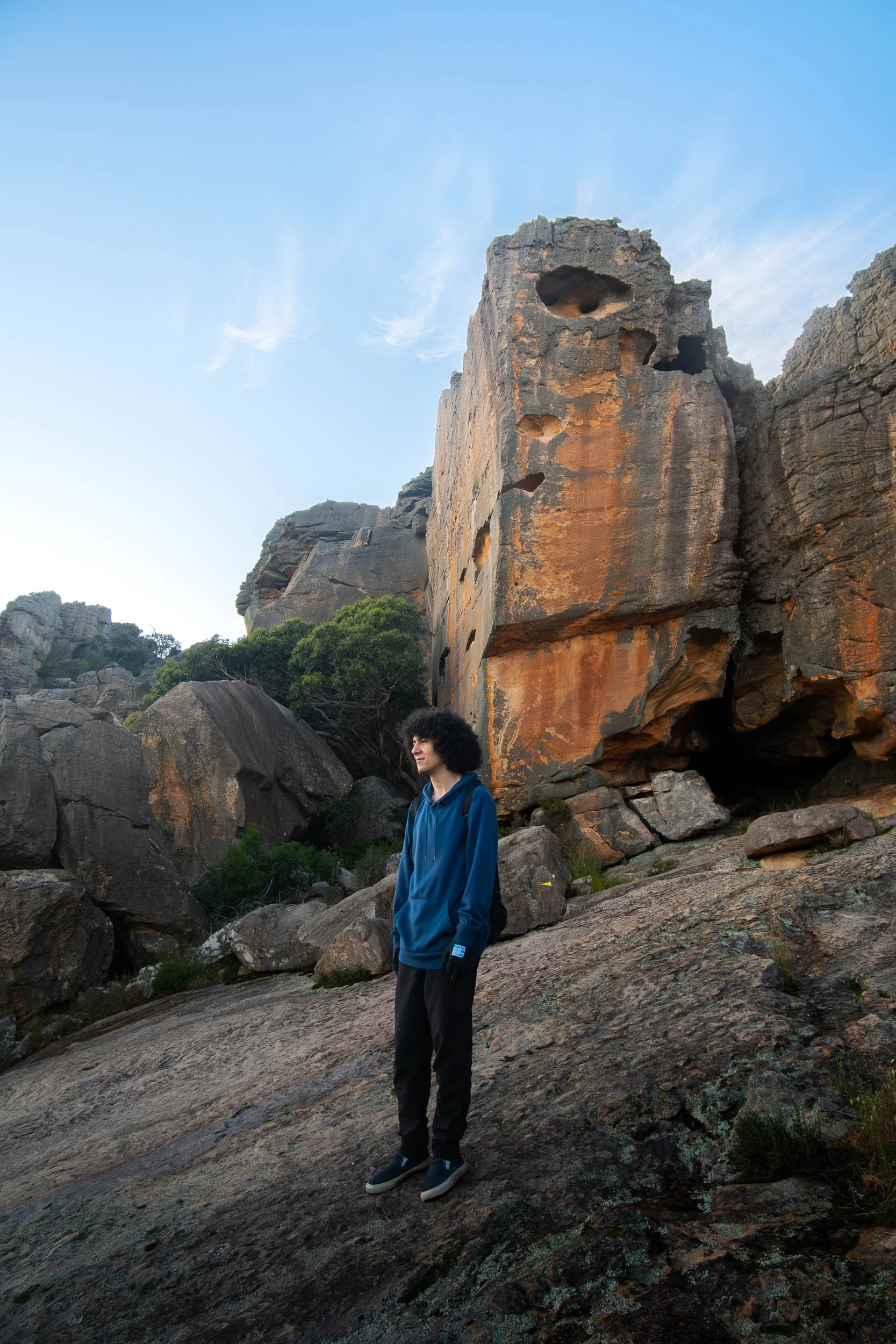 A person with curly hair in a blue hoodie stands on rocky terrain with large rocks and a cliff face with hollowed holes behind them.
