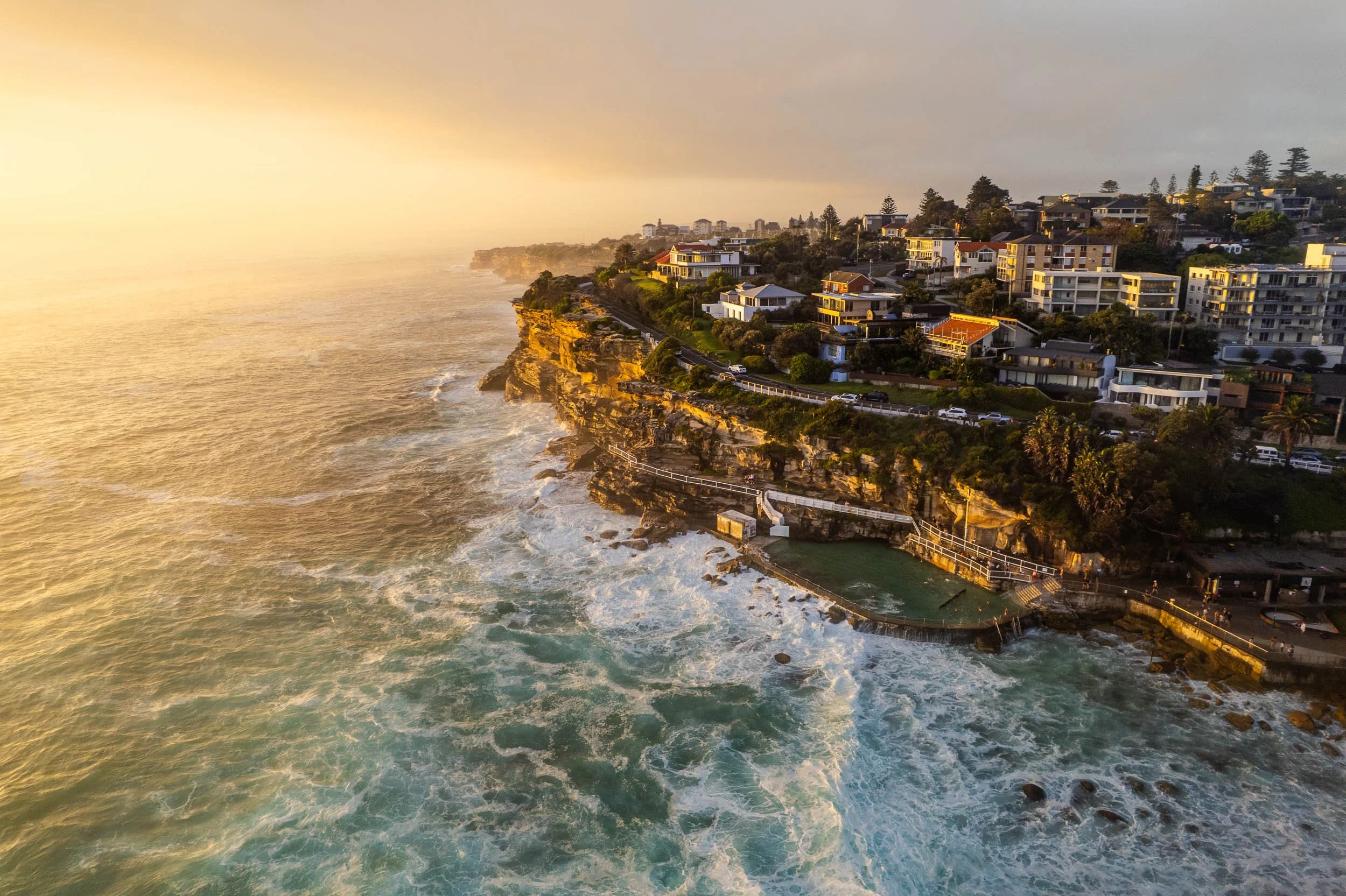 Aerial view of a coastal cliffside neighborhood at sunset, with houses overlooking the ocean and a rocky shoreline with crashing waves.