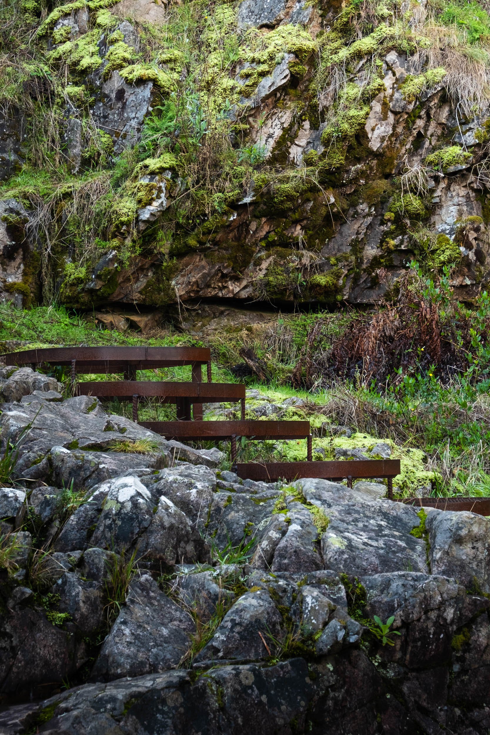 A set of rusted metal stairs ascending through a rocky, moss-covered outdoor landscape with dense greenery and a large mossy rock face.