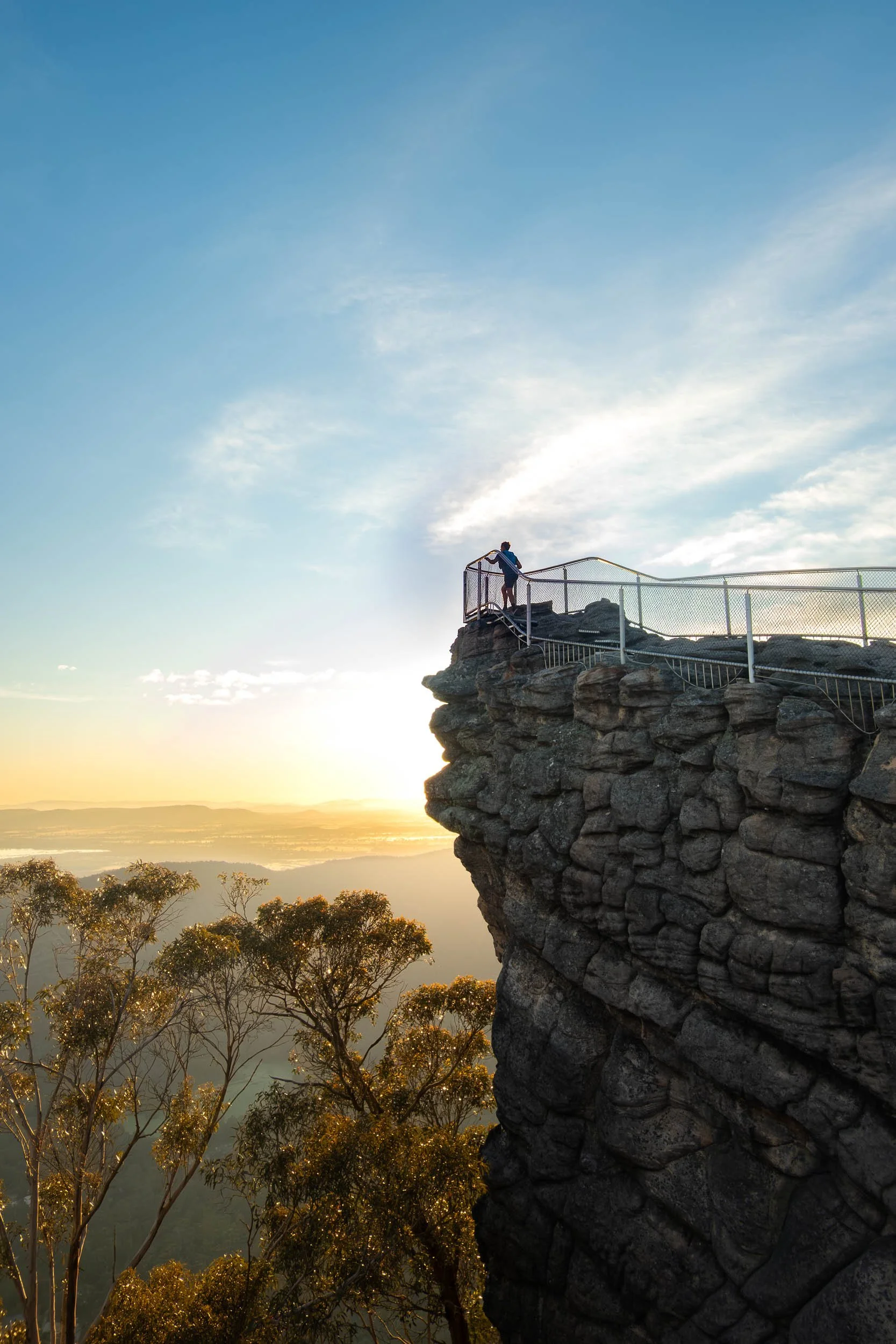 Person standing on a safety railing on a rocky cliff at sunset, overlooking a landscape with trees and water.