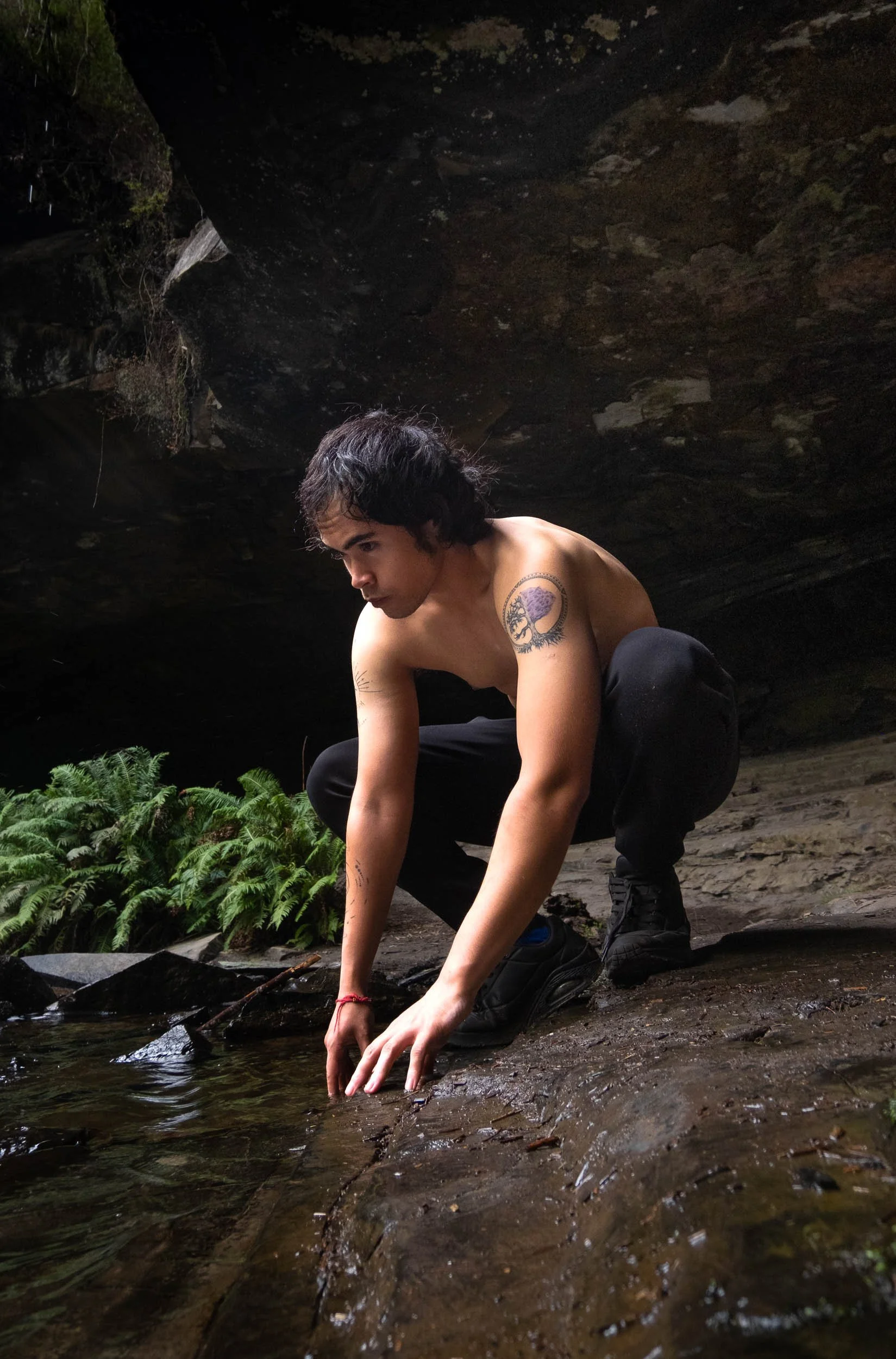 A shirtless man with tattoos crouches near a stream in a rocky, forested area.