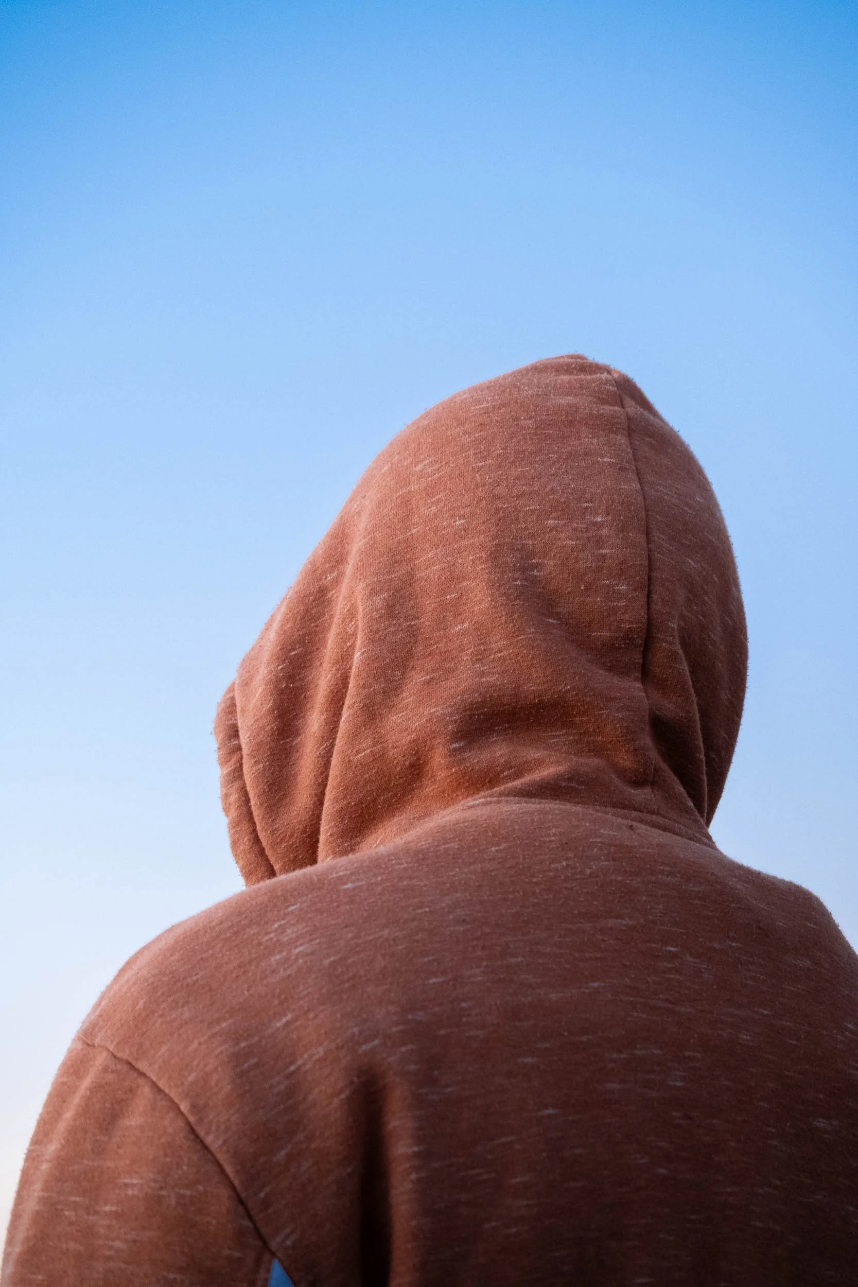 Back of a person wearing a brown hoodie, looking up at a clear blue sky.