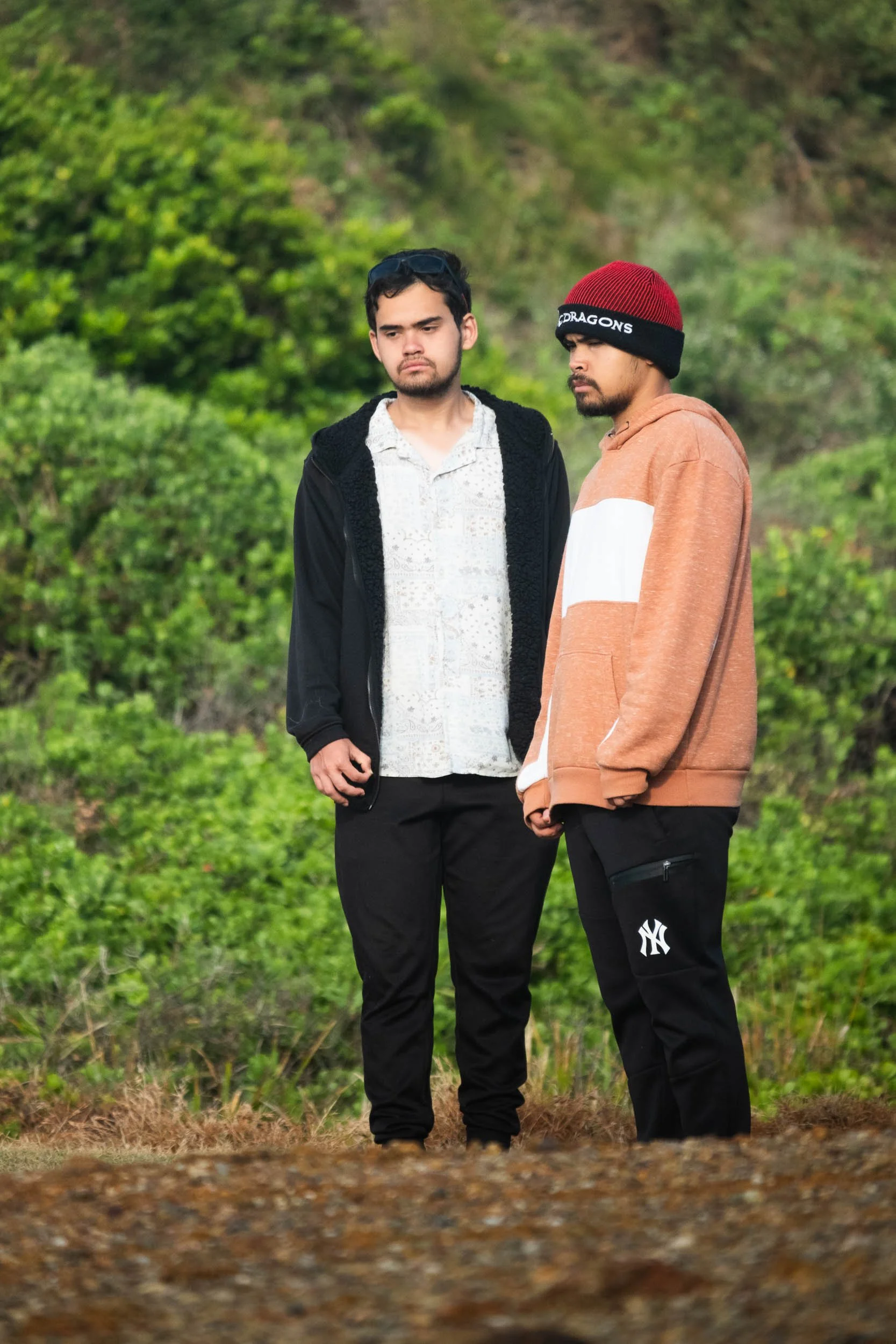 Two young men standing outdoors on a grassy hillside, dressed casually with serious expressions.