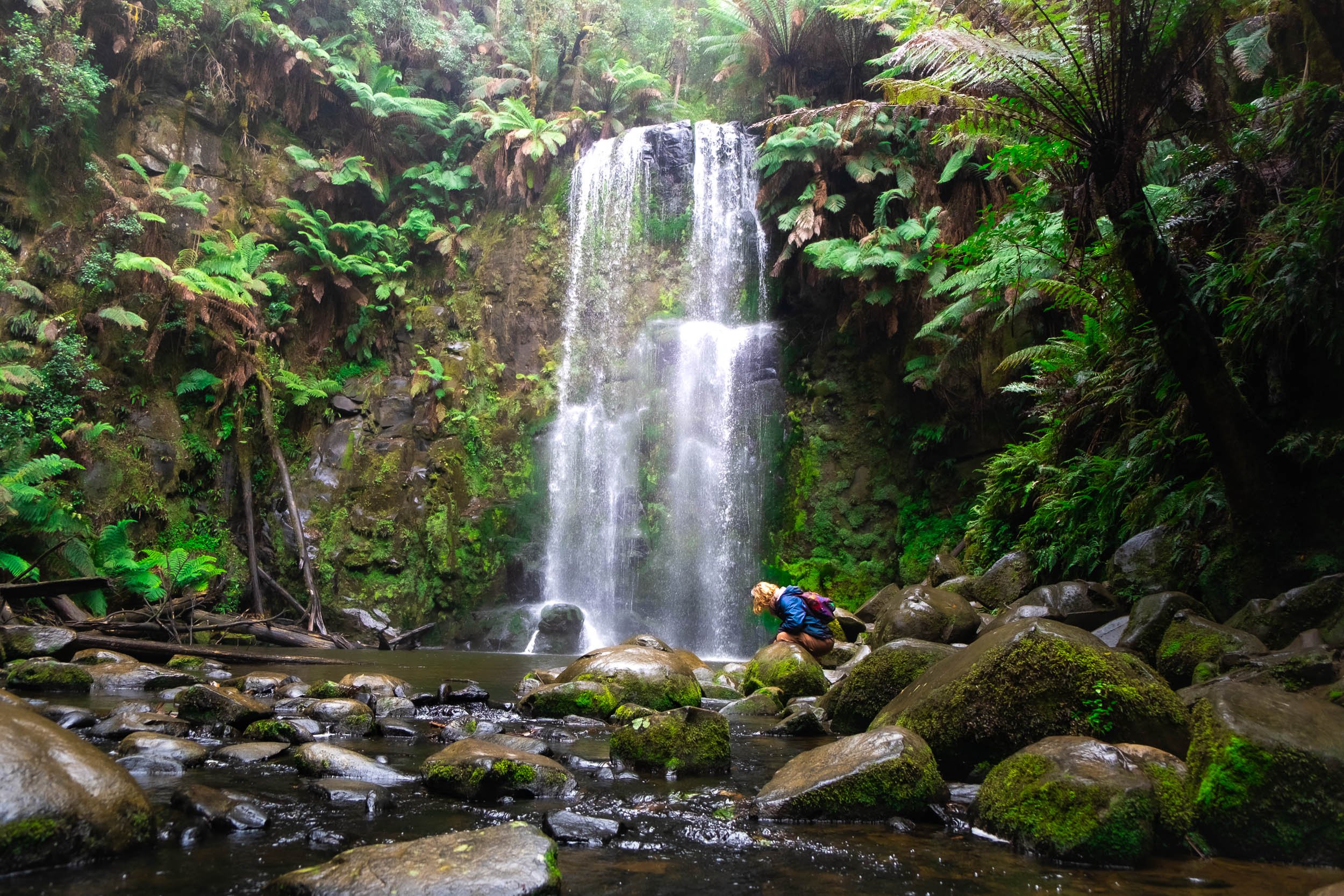 A person crouching on rocks in a lush green forest with a waterfall cascading in the background.