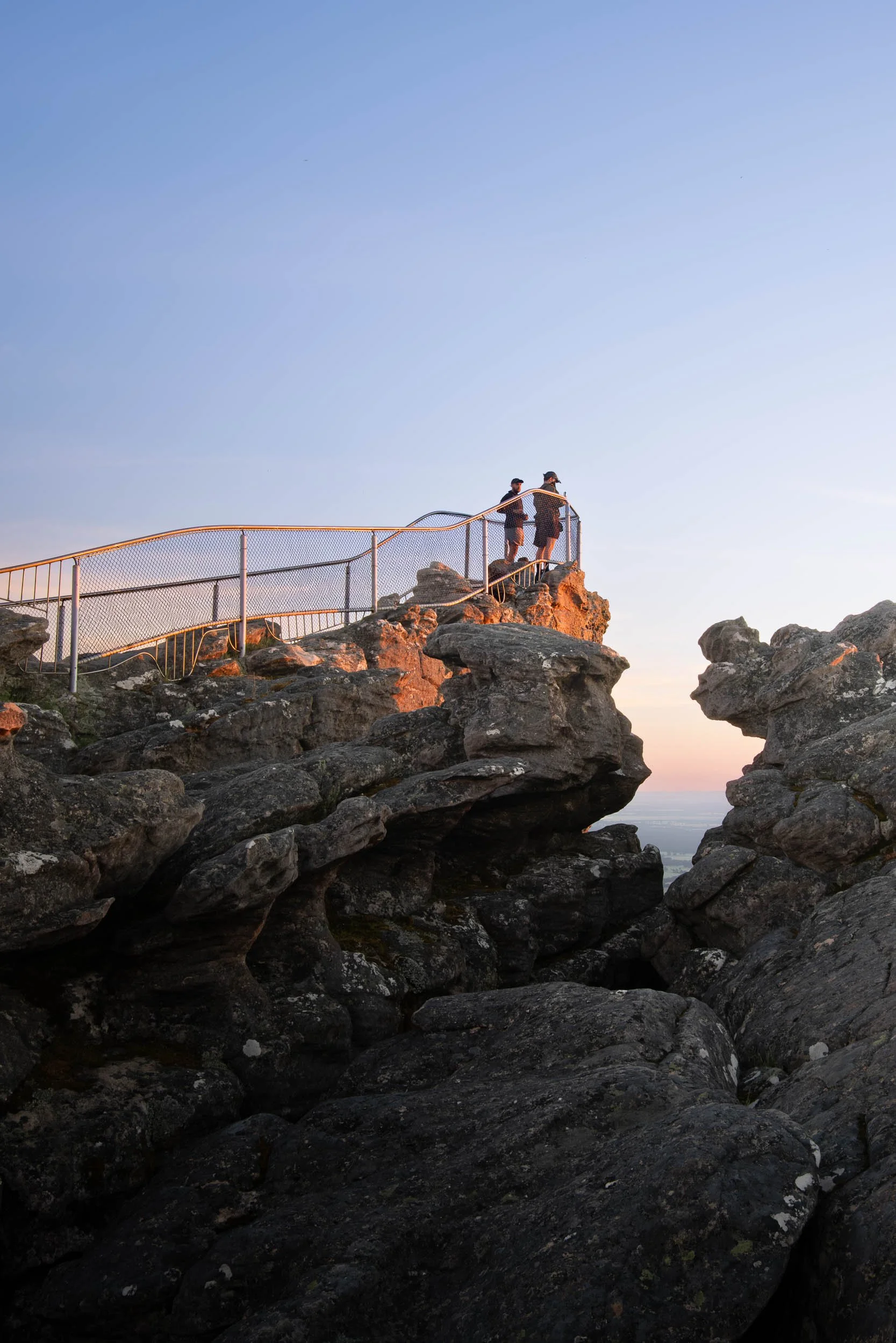 Two people standing on a rocky outcrop with a safety railing, during sunset, overlooking a landscape.