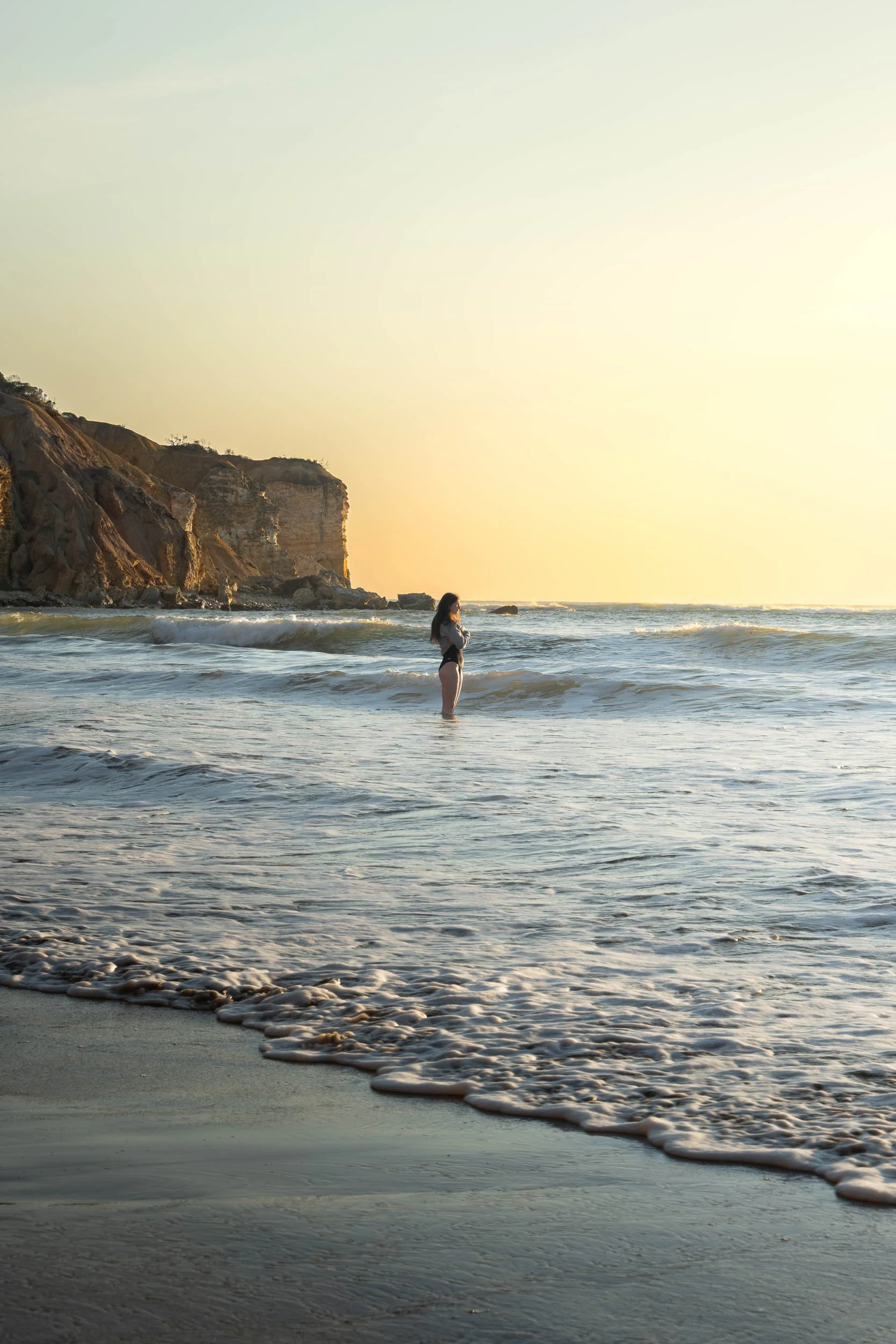 A woman standing in the shallow waters of a beach during sunset, with cliffs in the background.