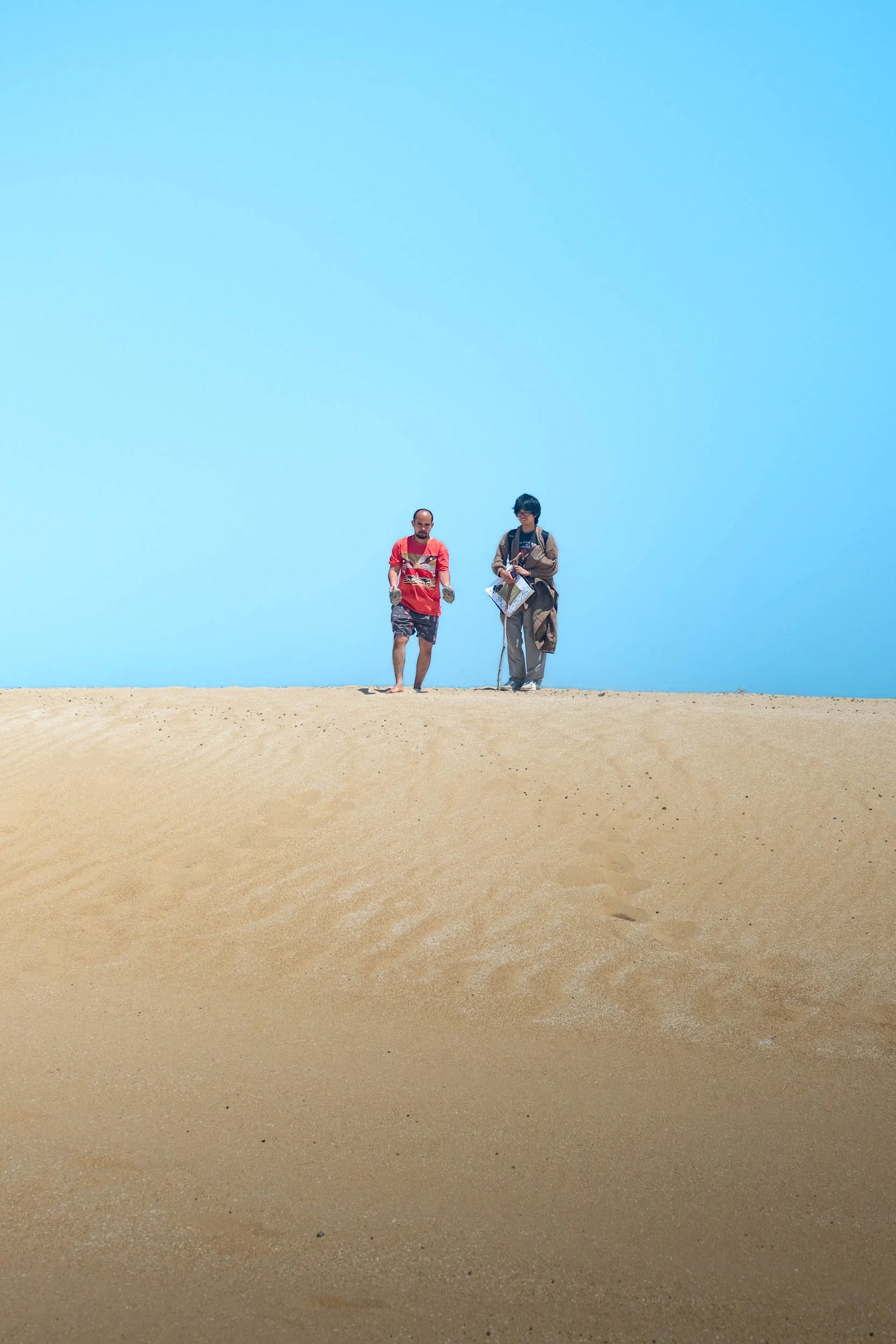 Two people walking on a sandy hill or dune with a clear blue sky in the background.