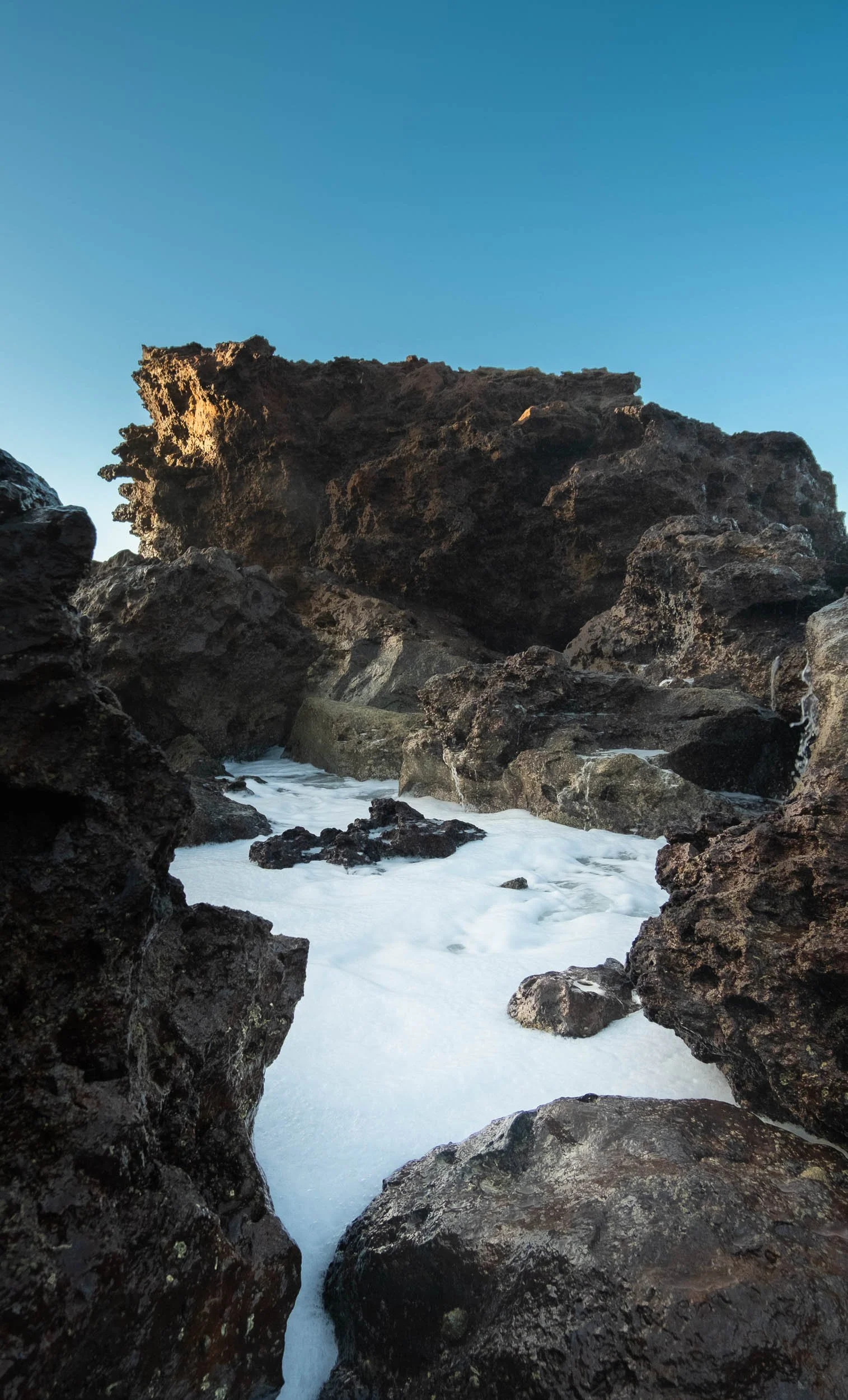 Rock formations along the coast with foamy waves in the foreground and a clear blue sky above.