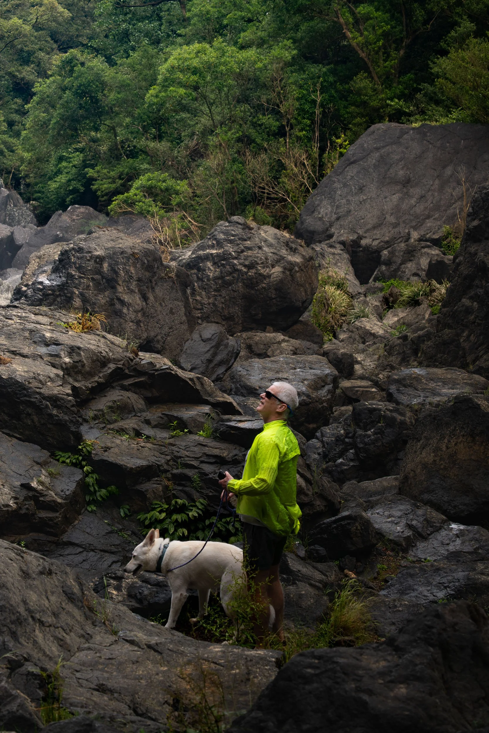 A man with gray hair wearing a bright green jacket and black shorts, holding a leash with a white dog, stands among large black rocks and green foliage in a forested area.