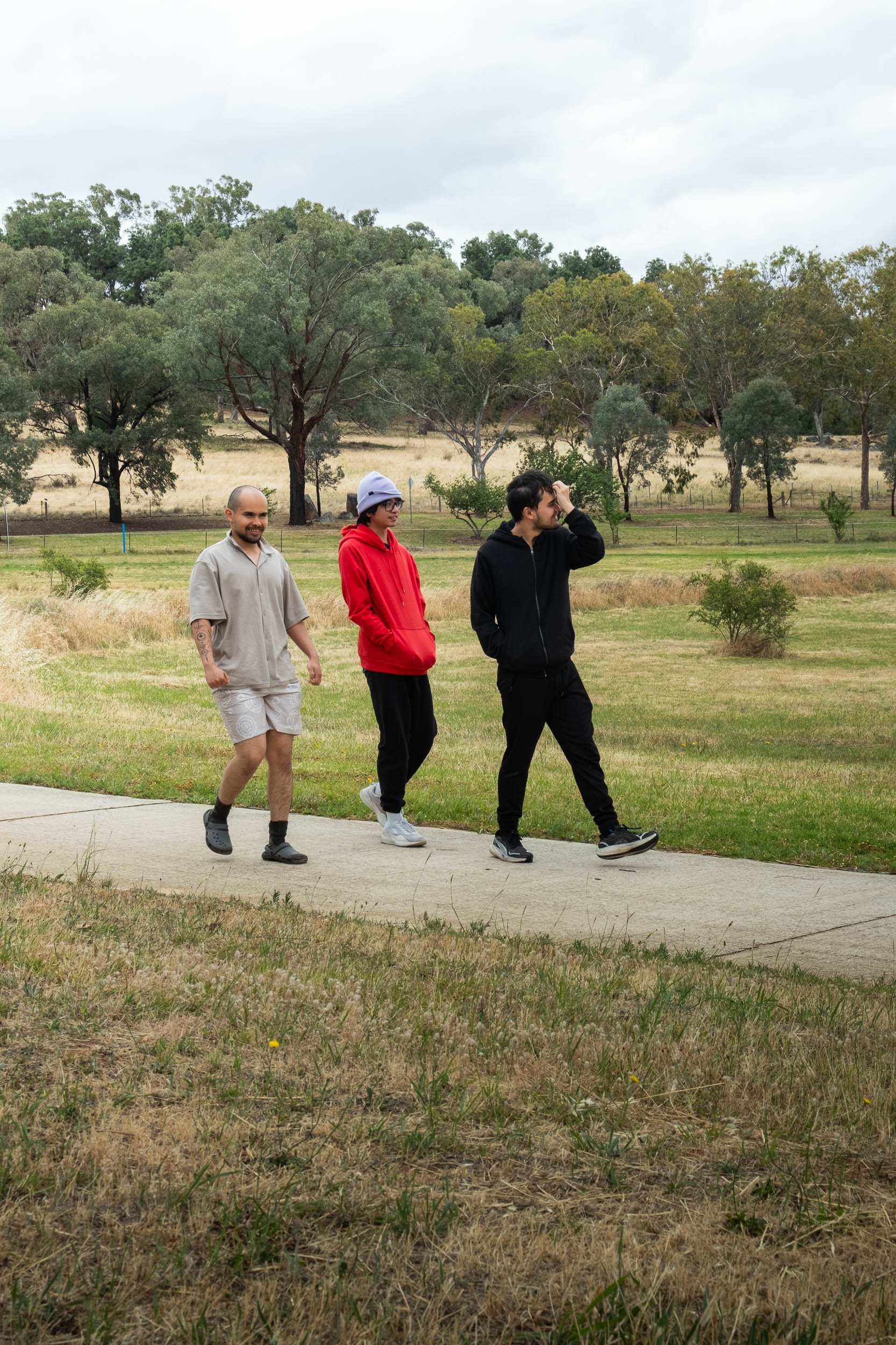 Three young men walking on a sidewalk in a park, with grassy fields and trees in the background on a cloudy day.