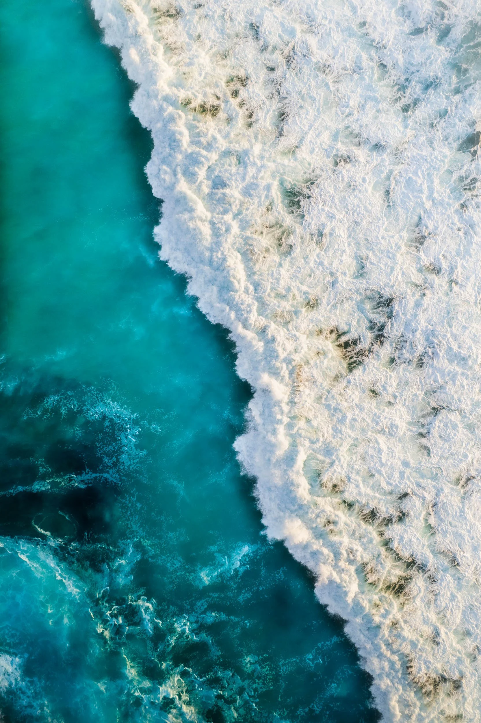 Aerial view of ocean waves crashing onto the shore, showing white foam and turquoise water.