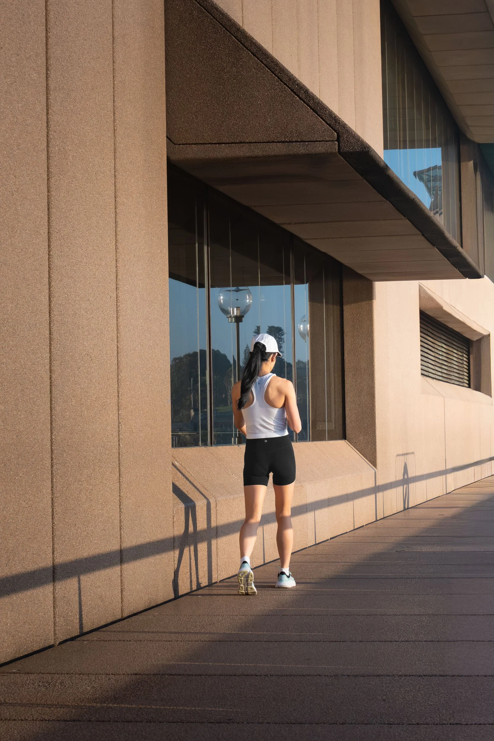 A woman dressed in white tank top and black shorts, wearing a white cap and athletic shoes, walking outside a modern building with large glass windows during sunset.
