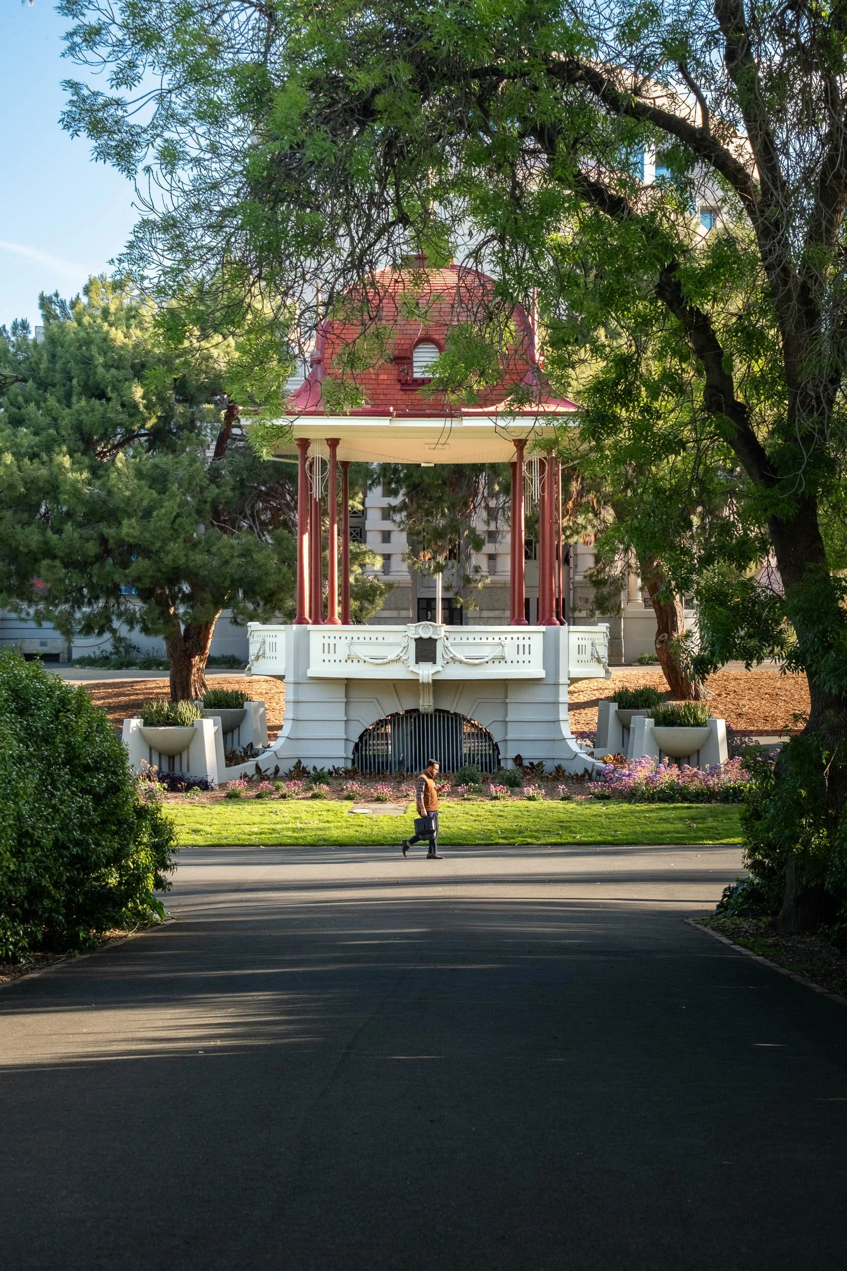A person walking past a decorative gazebo with a red roof in a park, surrounded by trees and greenery, during daylight.