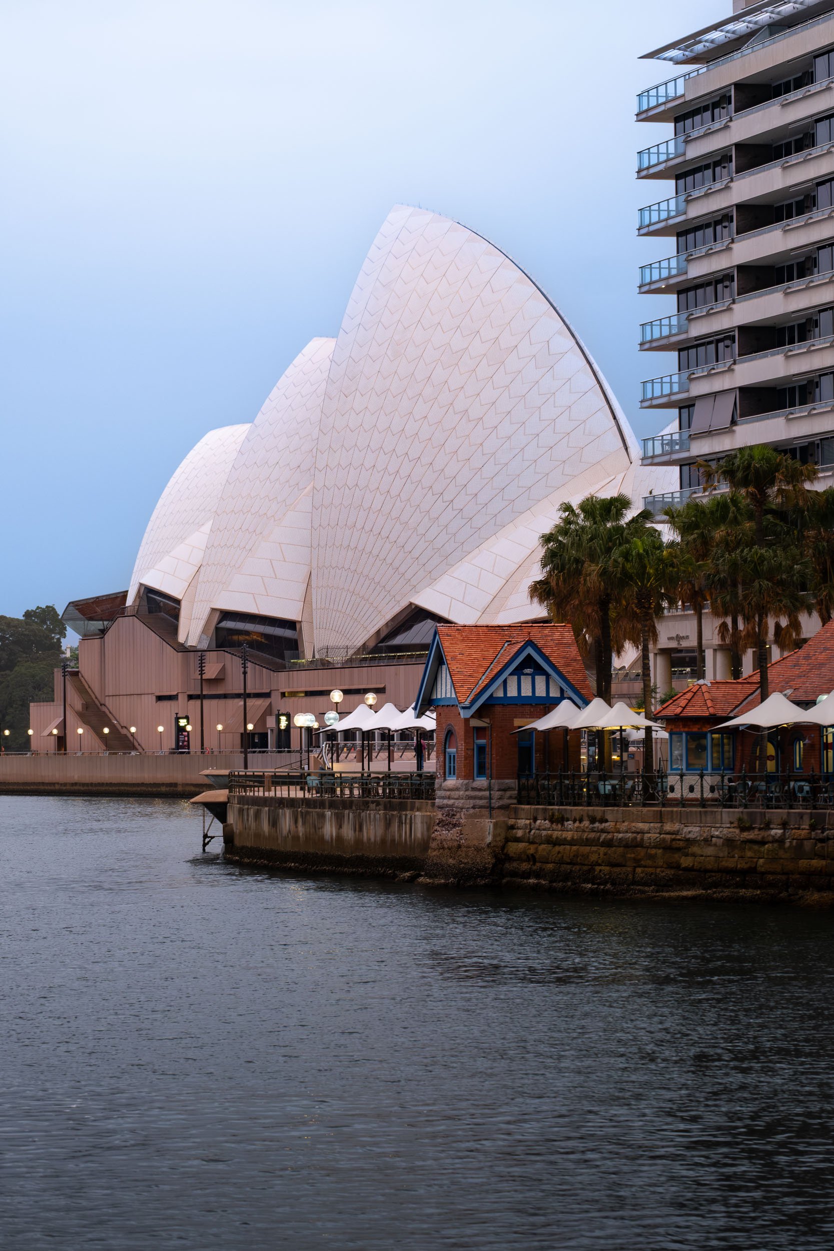 Sydney Opera House with surrounding waterfront, nearby high-rise building, and small colorful houses with outdoor seating, palm trees, and water in the foreground.