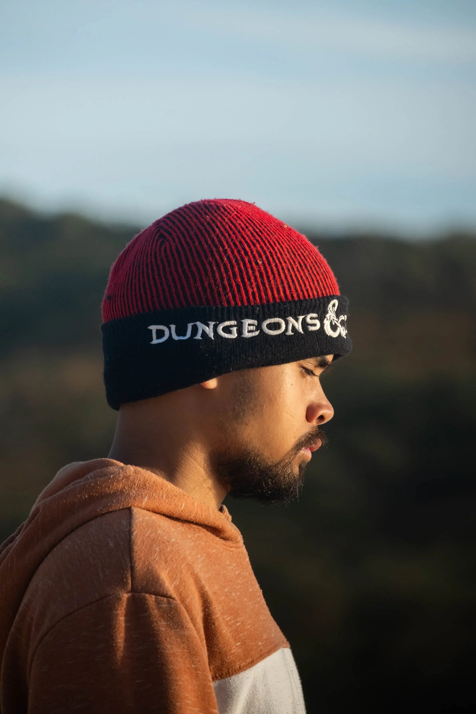 A man with a beard and closed eyes, wearing a red and black 'Dungeons' beanie, stands outdoors during sunset or sunrise with a blurred landscape in the background.