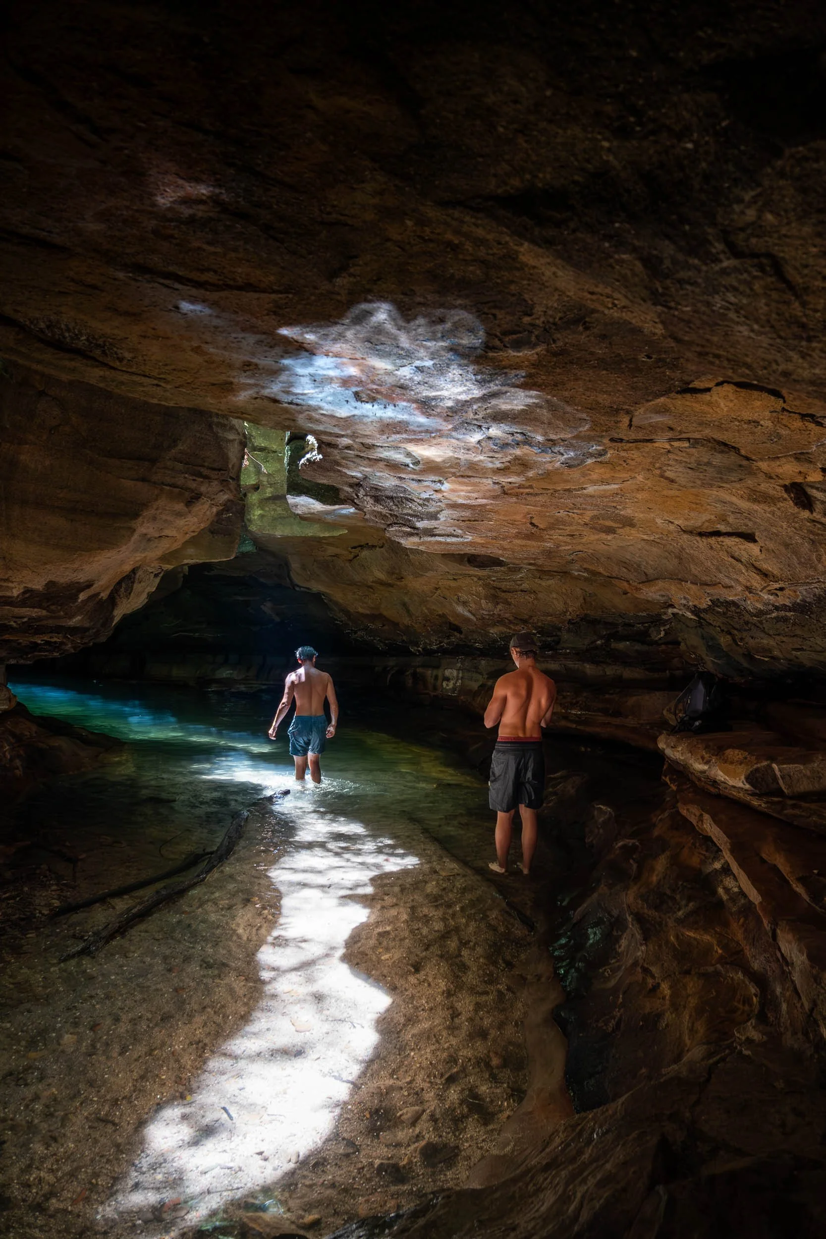 Two shirtless men in shorts wading through a shallow pool inside a dark cave.