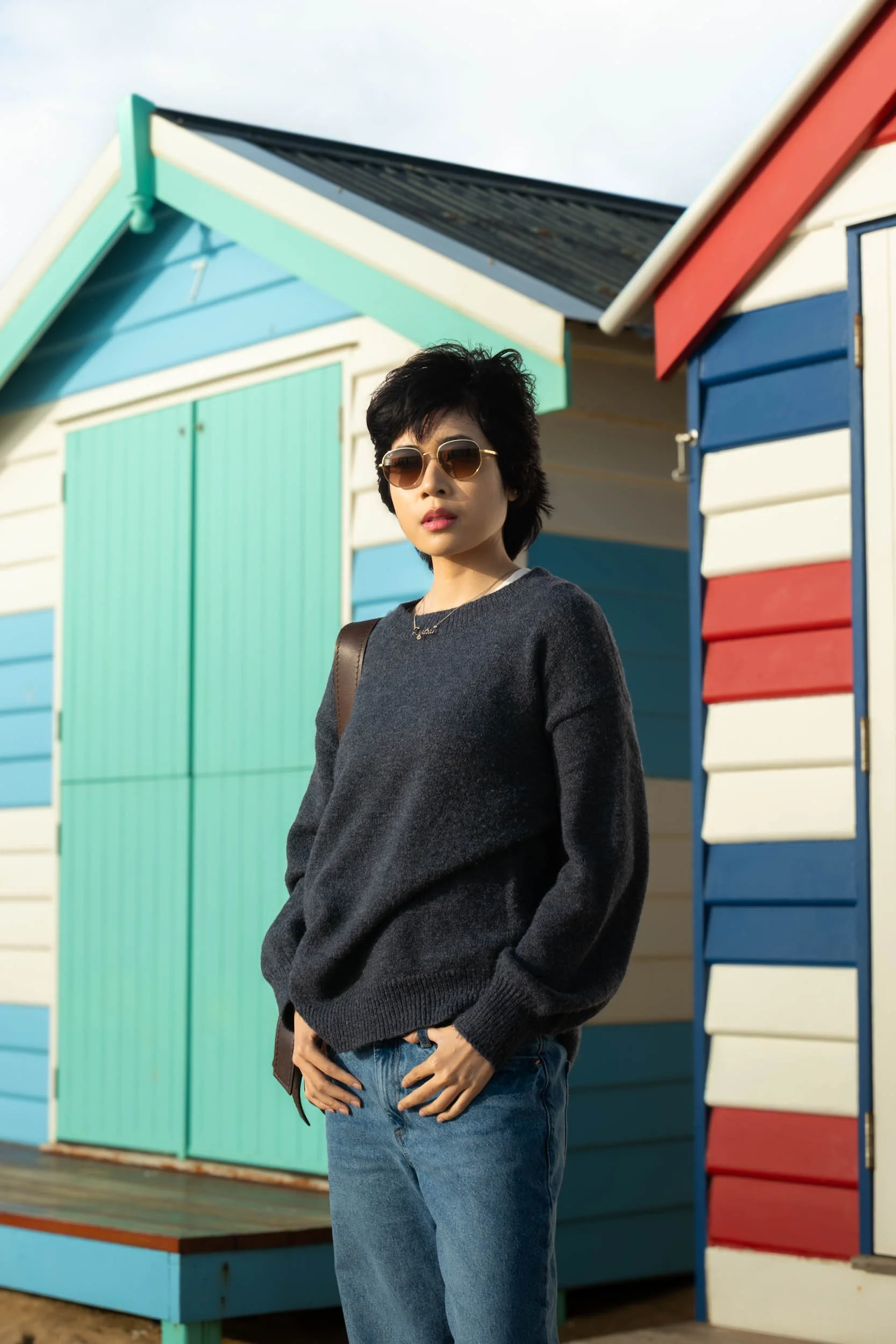 Young woman with short black hair wearing sunglasses, a dark gray sweater, and blue jeans standing in front of colorful beach huts during daytime.