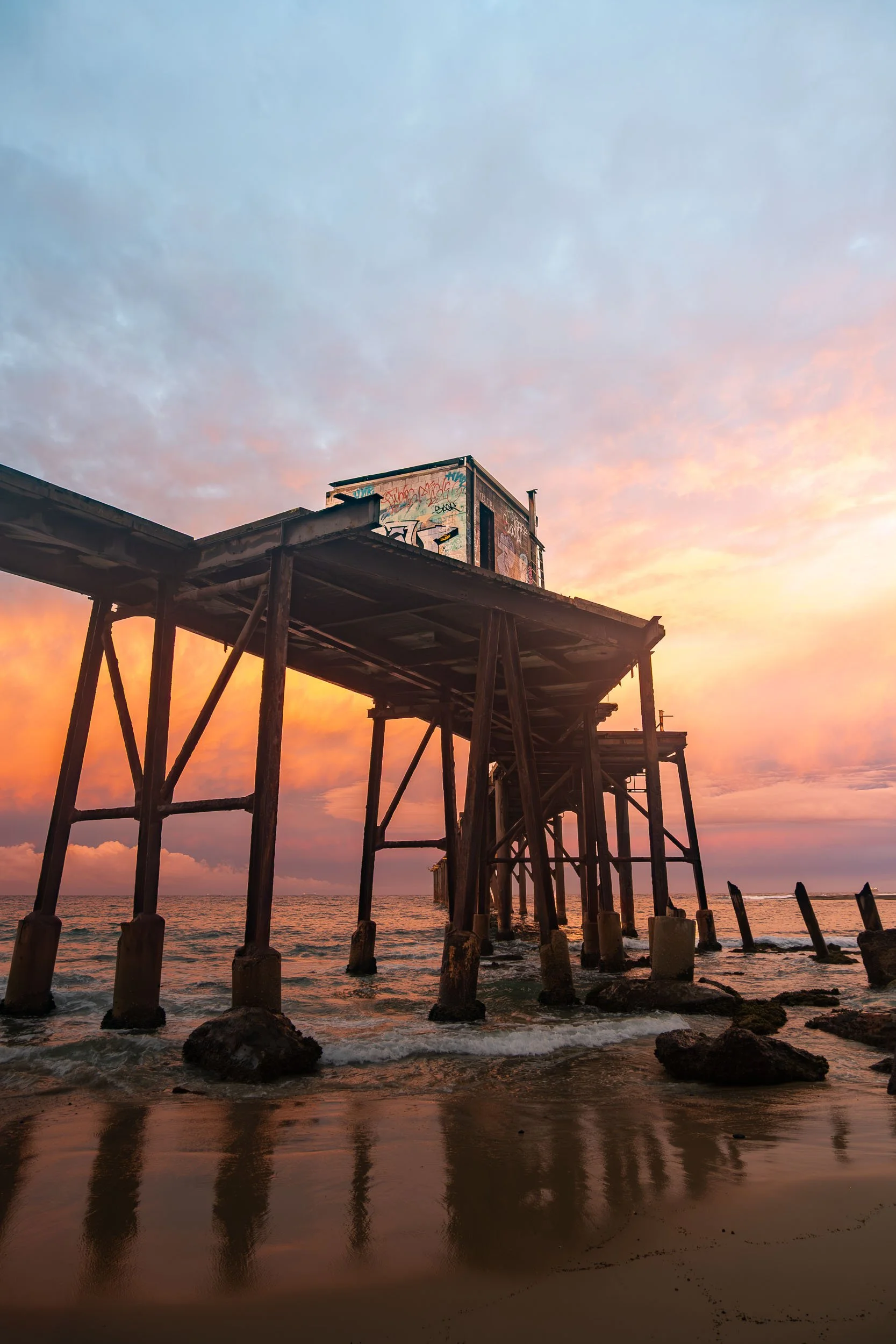 An abandoned pier with a small building on top, viewed during sunset with pink and orange clouds reflected on wet sand.