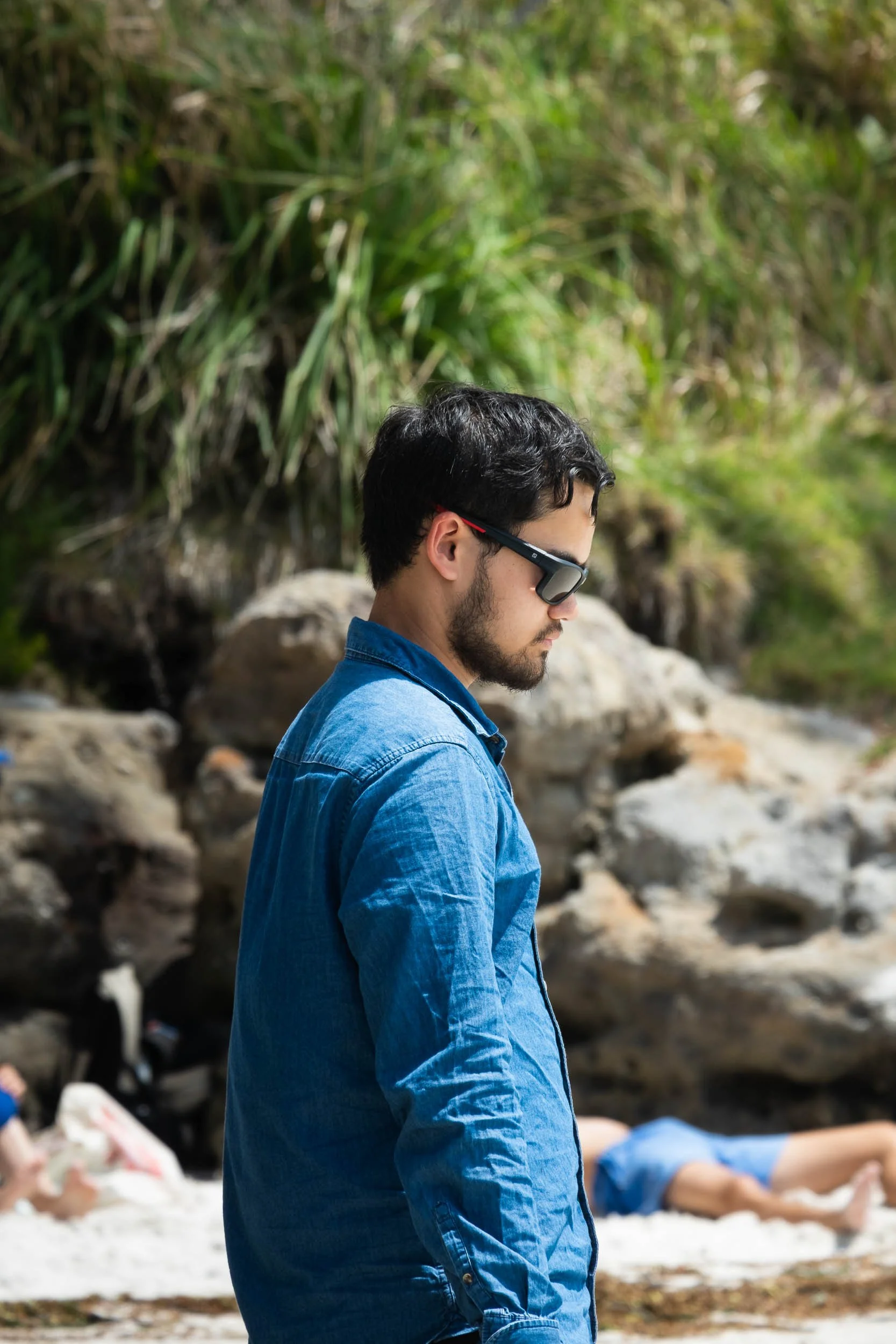 A man with dark hair, beard, wearing sunglasses and a blue denim shirt, stands outdoors near a rocky area with green foliage in the background.