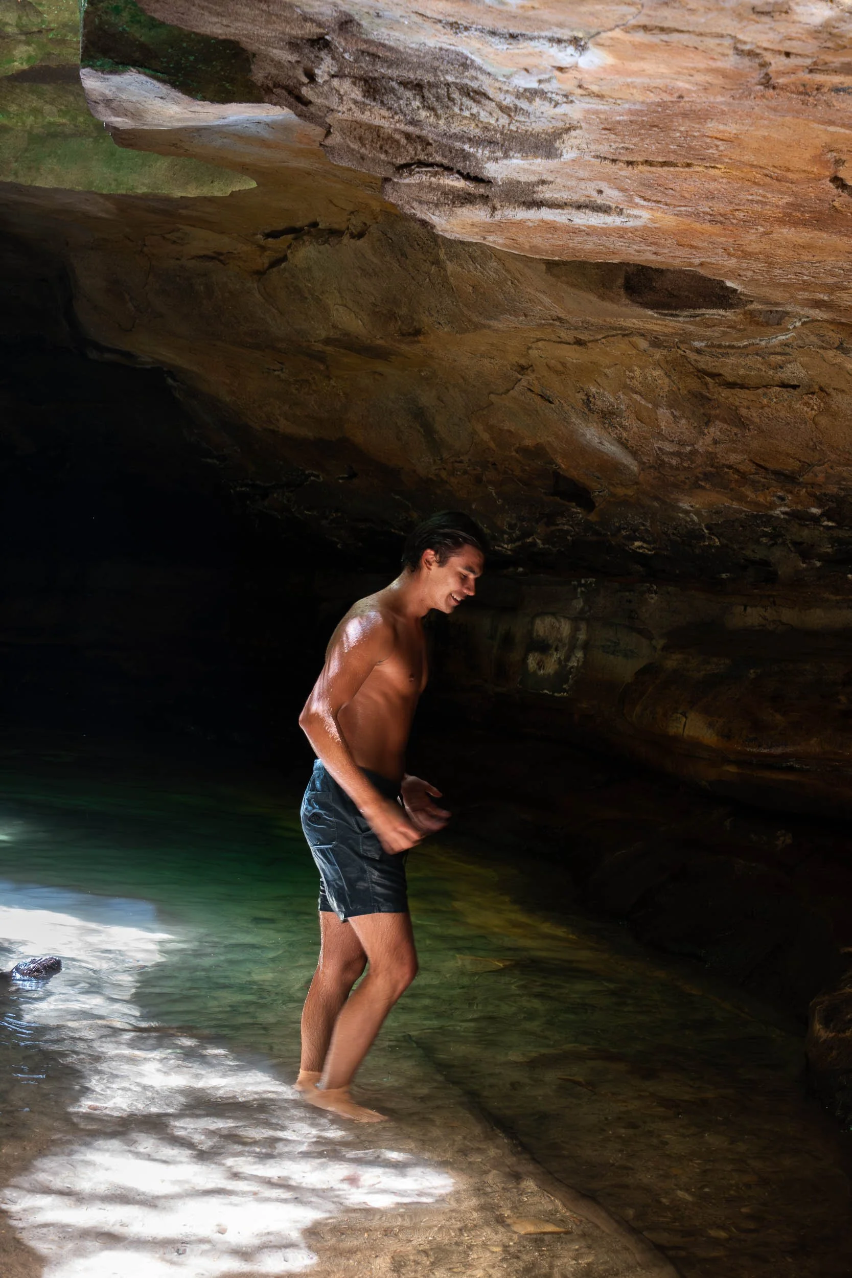 A young man in black swim shorts enjoying the water in a rocky cave, with a large rock ceiling above and shallow water around his feet.