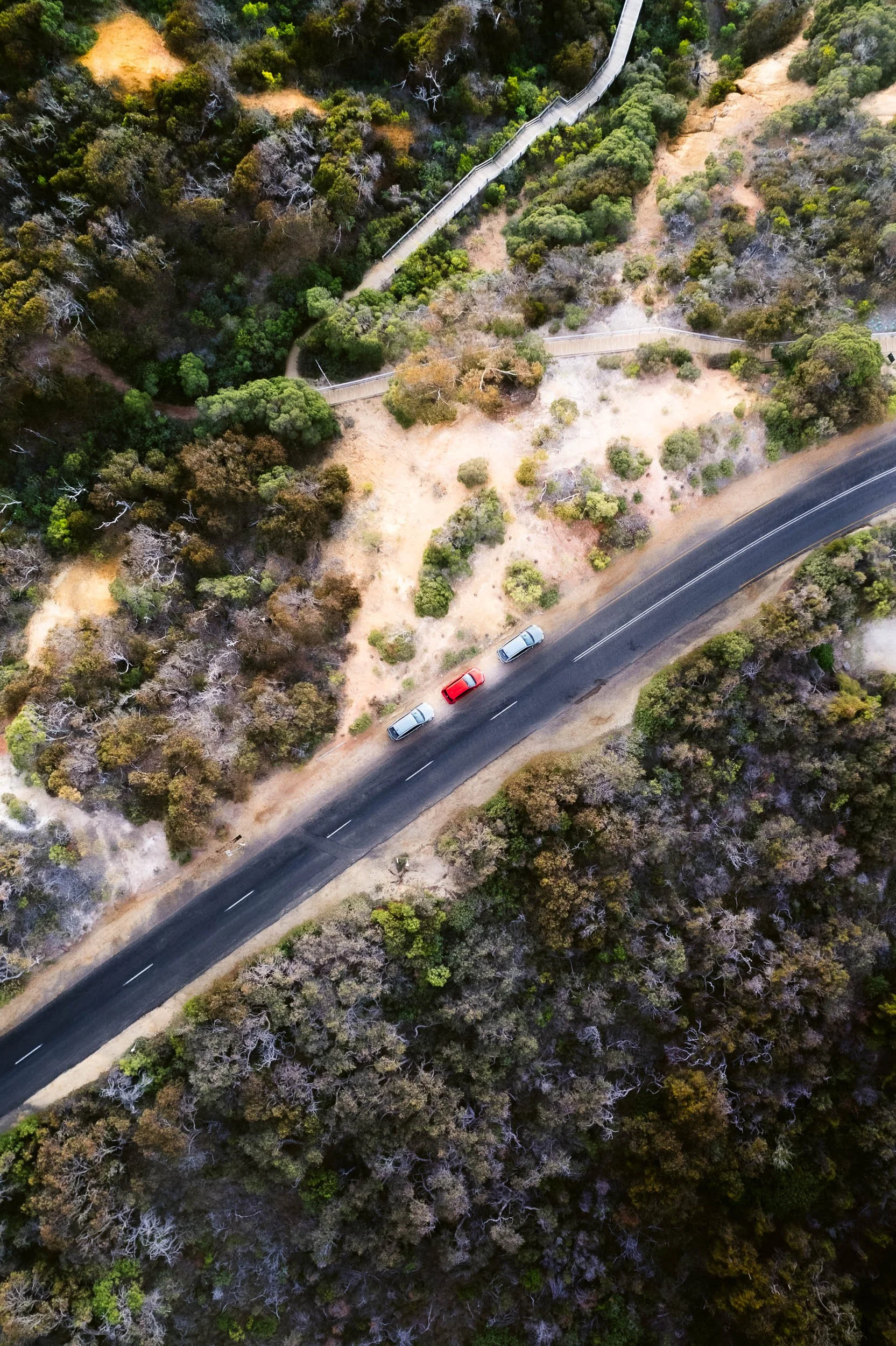 An aerial view of a winding two-lane road through a wooded area with three cars traveling on it, surrounded by green and brown trees and shrubs.
