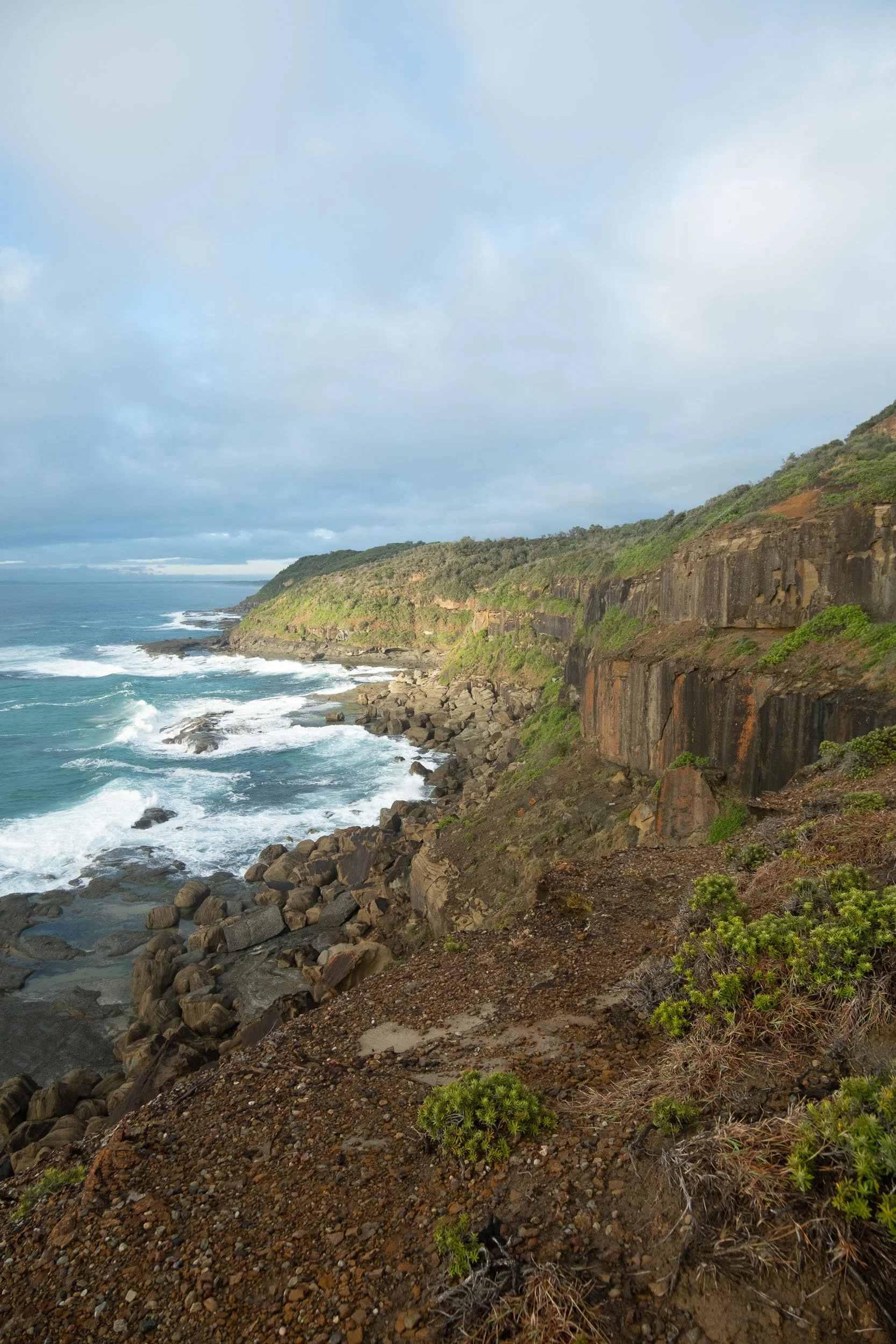 Coastal cliffs with green vegetation, rocky shoreline, and waves crashing onto rocks, under a cloudy sky.