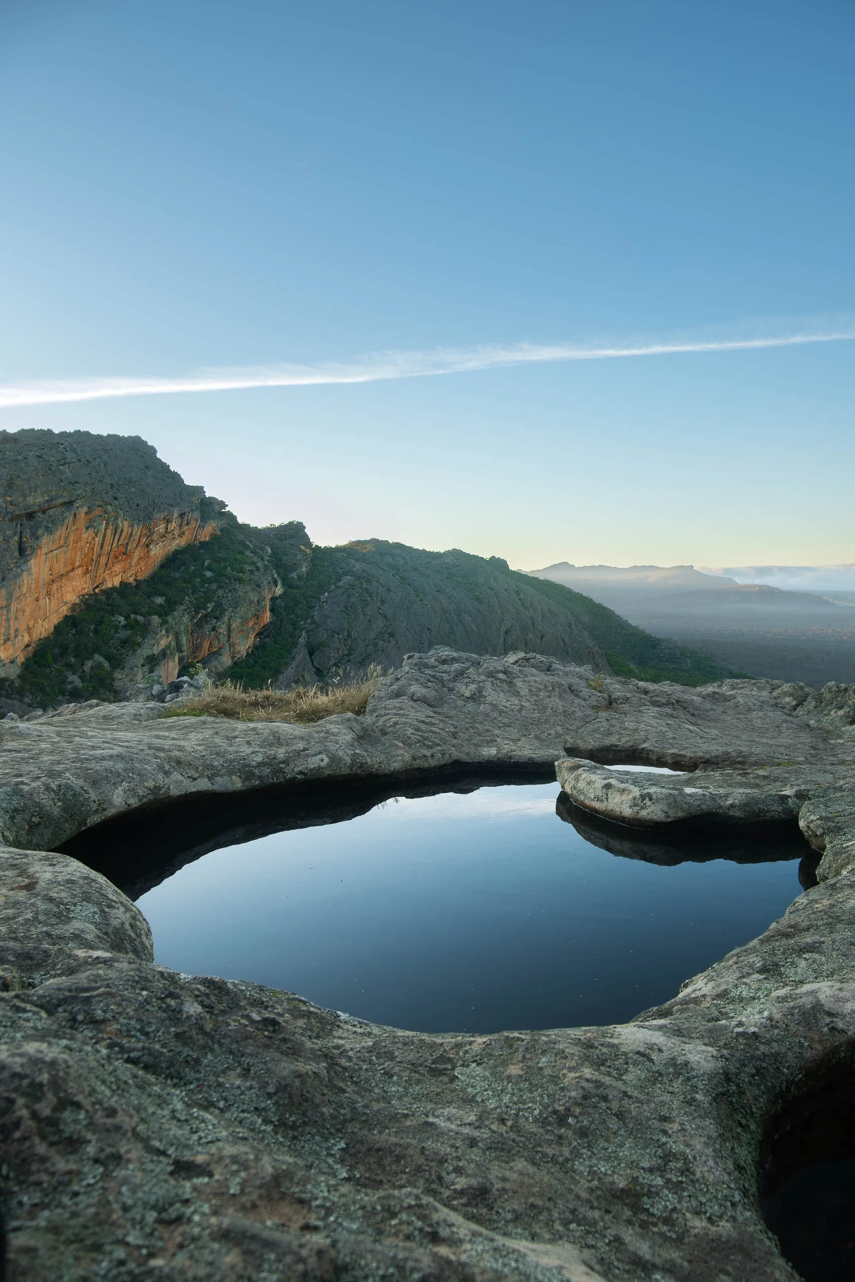 A landscape view of rocky terrain with a small pool of water, overlooking a valley and mountains under a clear blue sky.