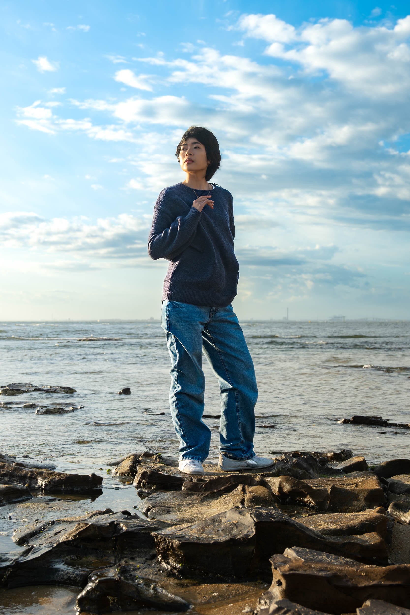 A woman standing on rocks by the ocean, wearing a dark sweater and jeans, with a cloudy sky in the background.