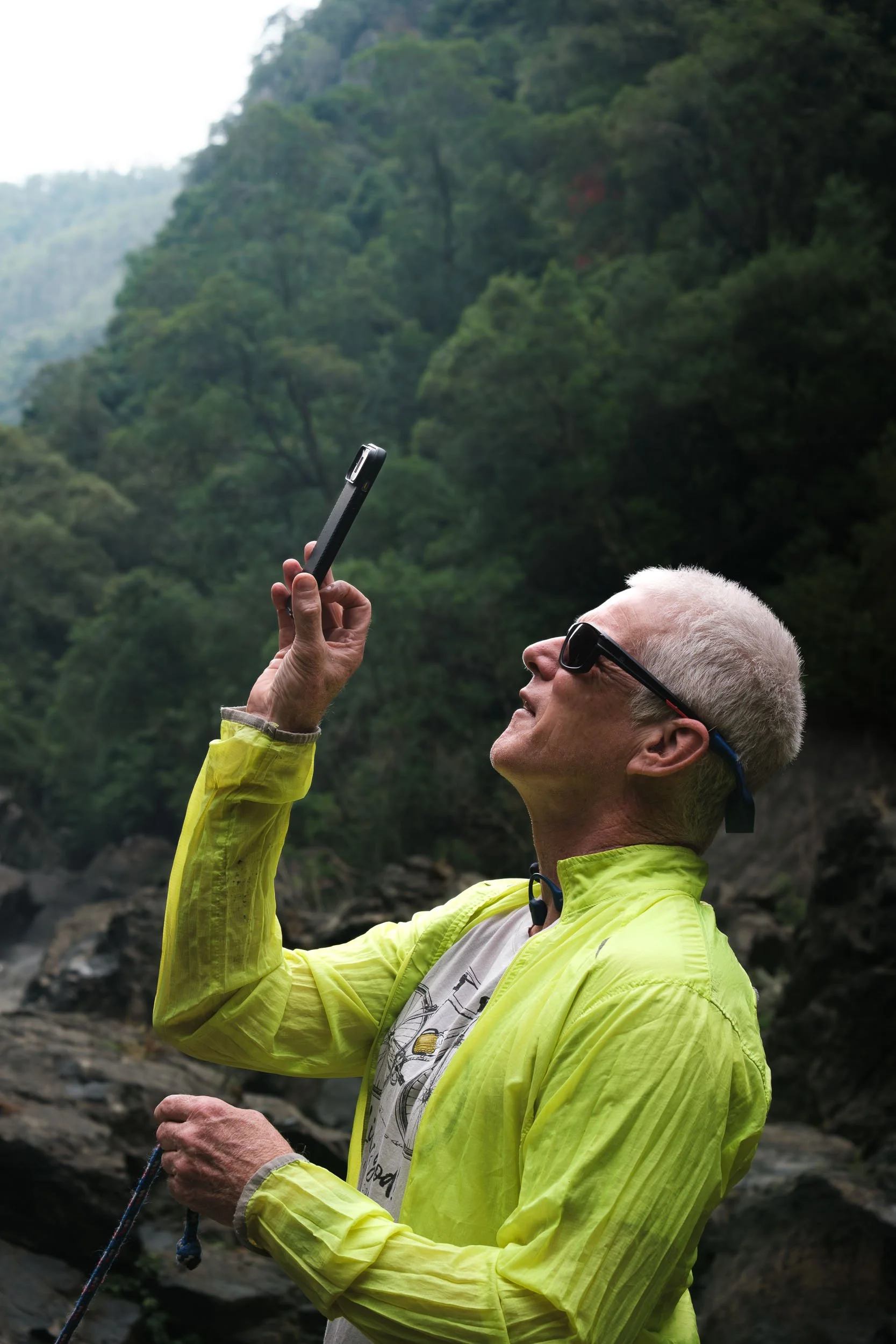 Older man with white hair and sunglasses taking a photo with a smartphone in a forested outdoor setting.