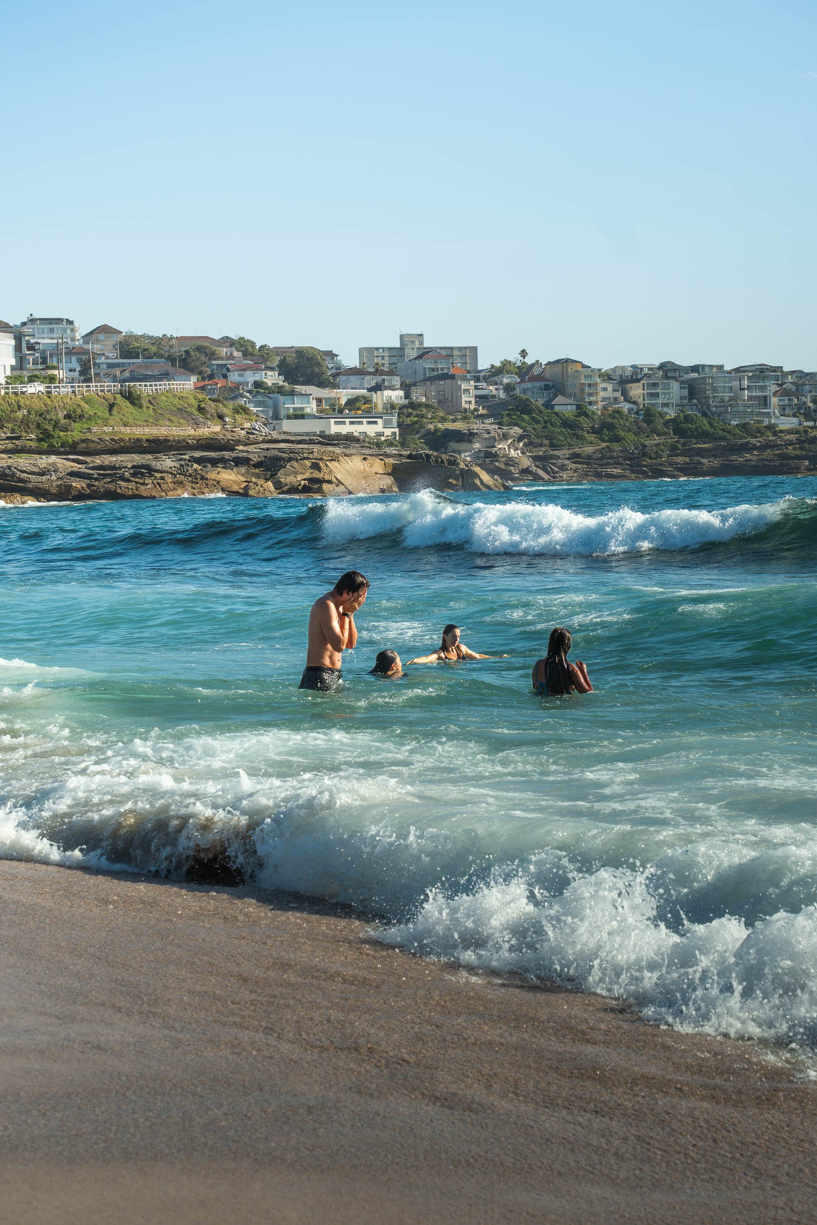 People enjoying a day at the beach, wading in the ocean with houses on a coastal hill in the background.