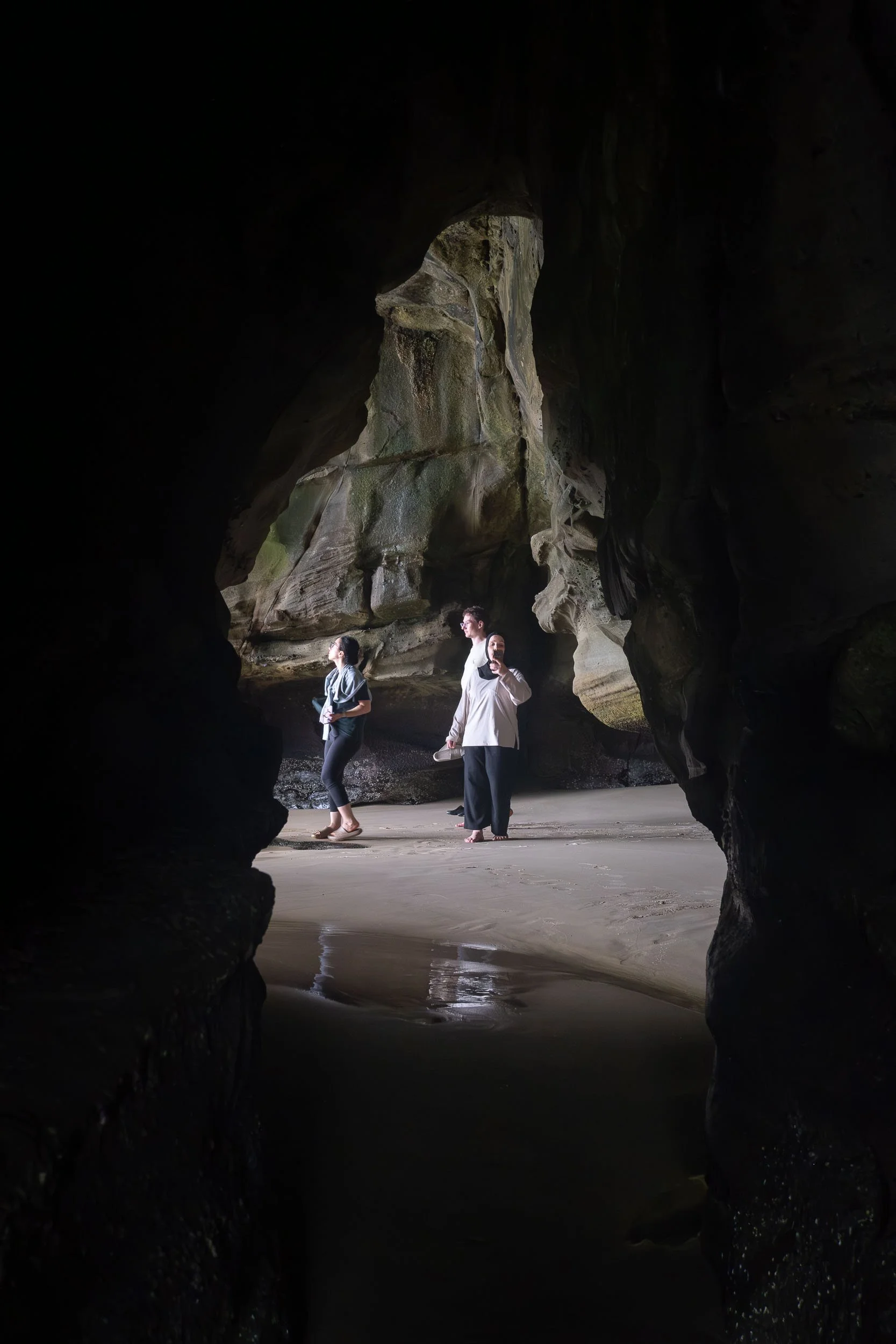 View of three people exploring a cave, seen through a dark rocky opening, with large cave walls and sandy floor visible.