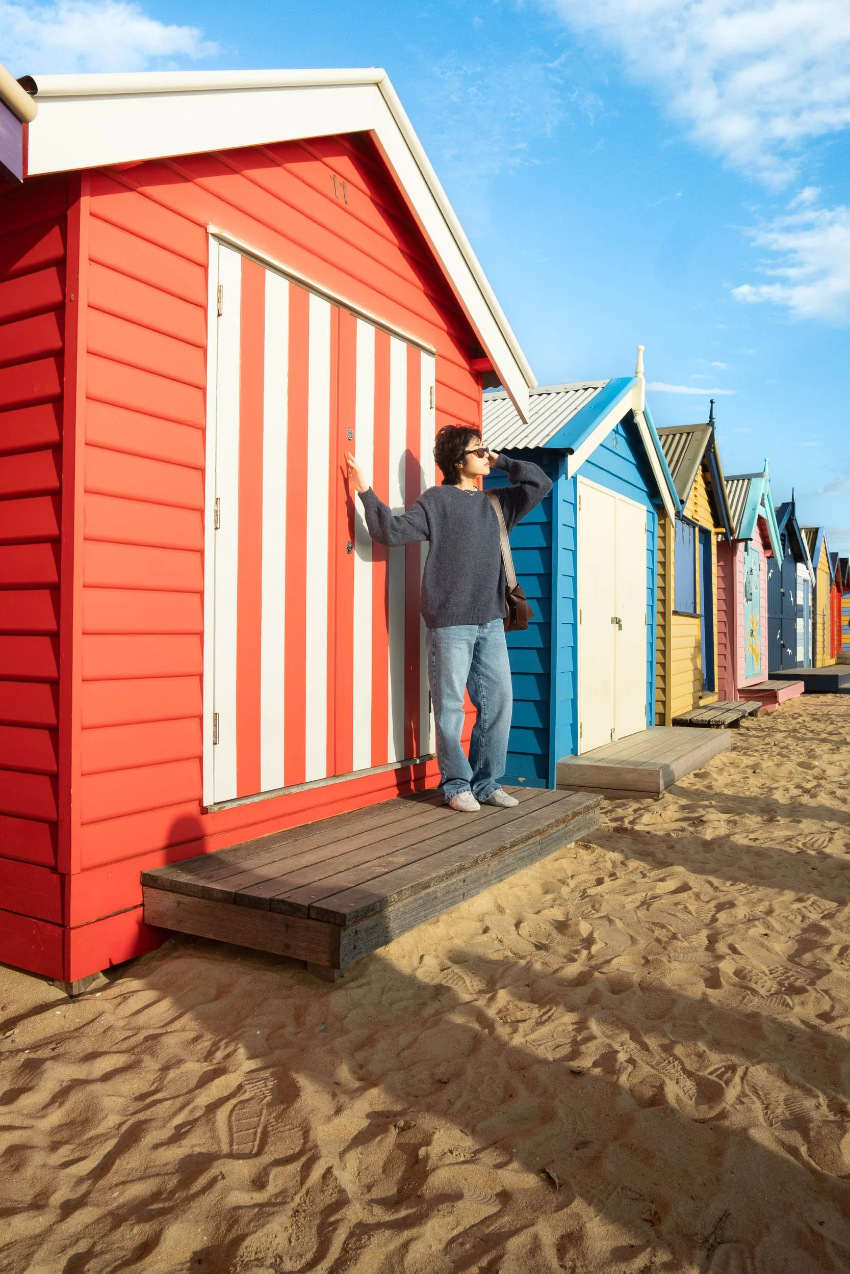 A woman standing on a wooden platform in front of a colorful beach hut with red and white vertical stripes. The beach huts are arranged in a line on sandy ground under a blue sky.