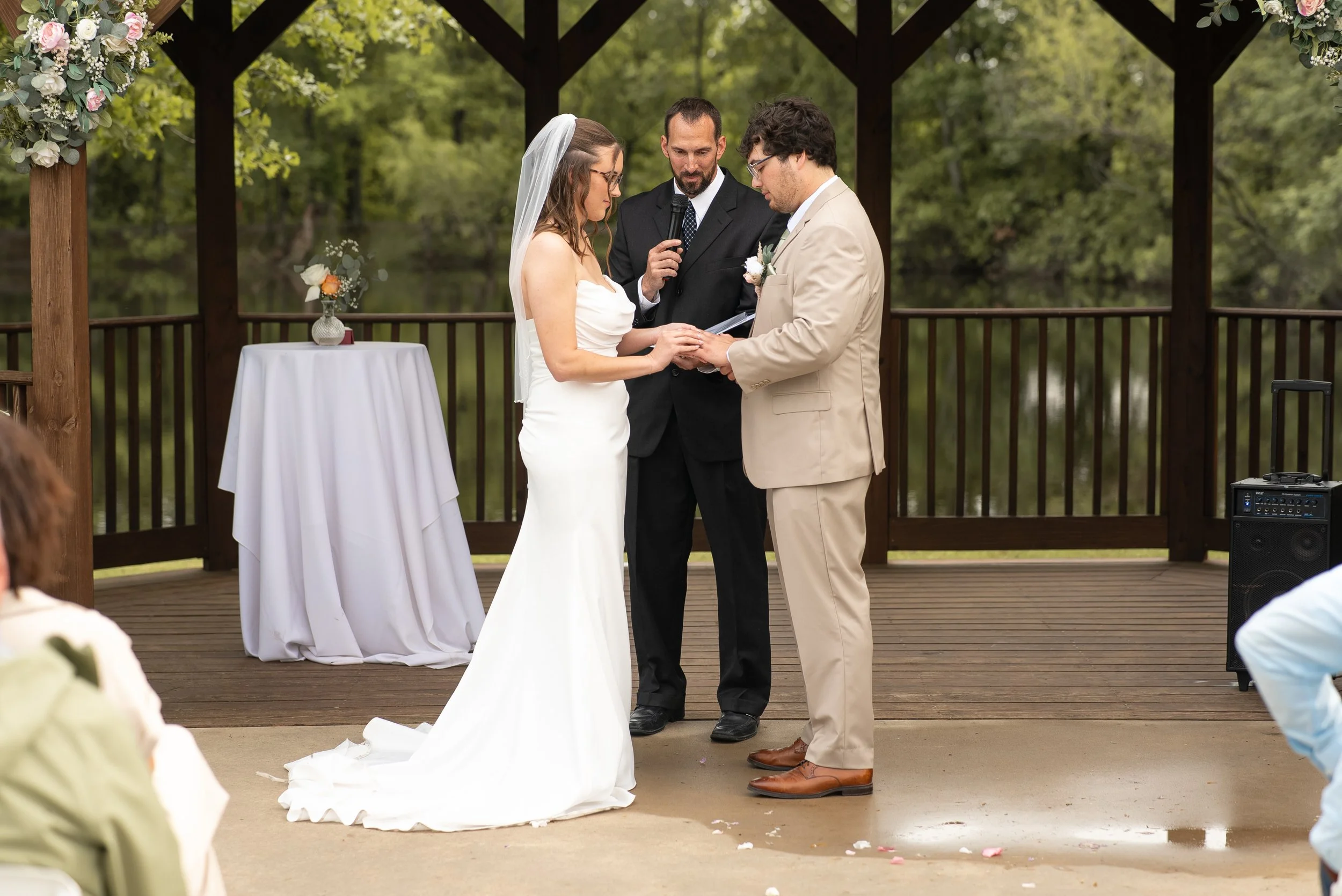 A couple exchanging wedding vows outdoors on a pavilion, with an officiant standing behind them, during a wedding ceremony surrounded by trees.