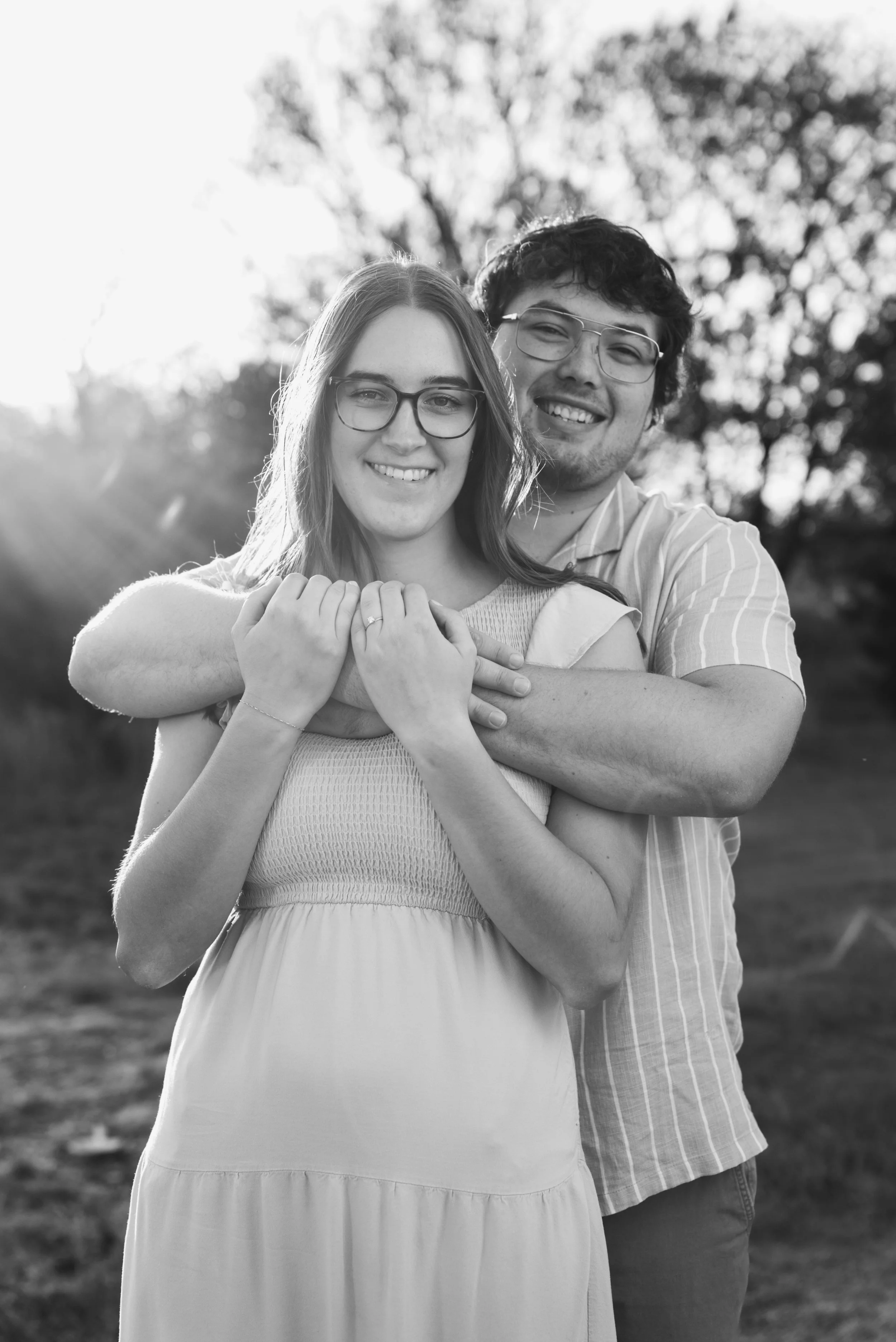 A young woman and young man are smiling and embracing outdoors, with the man standing behind the woman and wrapping his arms around her shoulders. They are wearing glasses and dressed casually, with trees and sunlight in the background.