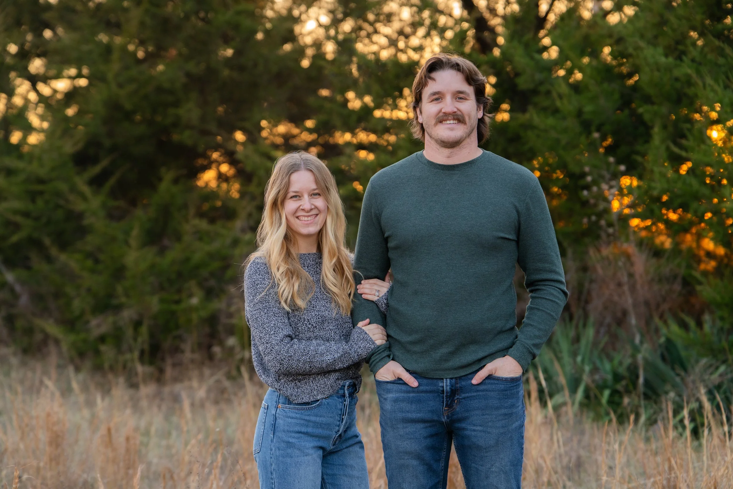 A smiling woman with blonde hair wearing a gray sweater and jeans standing next to a taller man with brown hair and a mustache, wearing a dark green sweater and jeans, outdoors during sunset with trees in the background.
