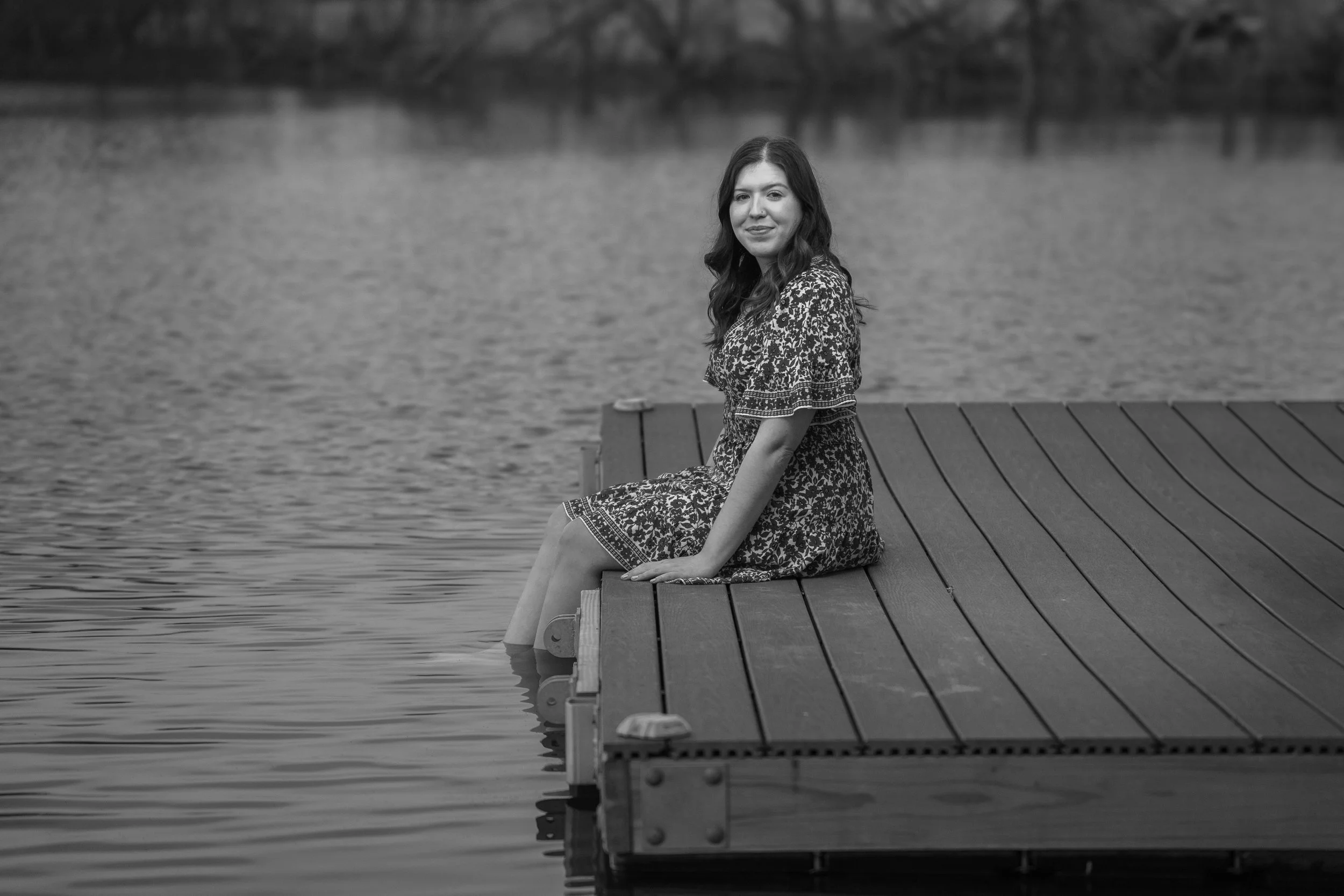A woman in a patterned dress sitting on the edge of a wooden dock with her feet in the water, by a lake or river, in black and white.