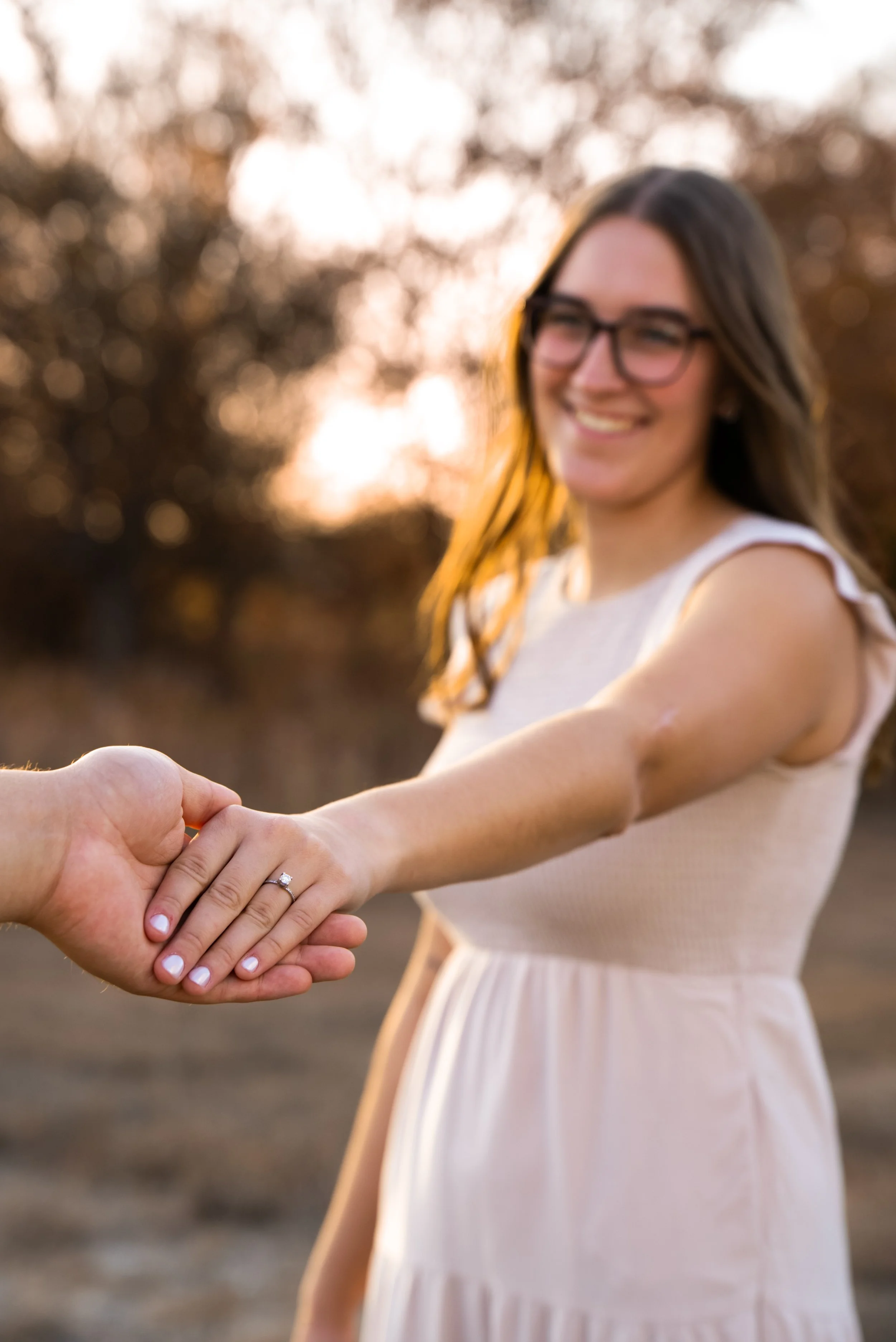 A woman with glasses and long hair smiling at the camera, holding hands with someone, showing an engagement ring on her finger during sunset outdoors.