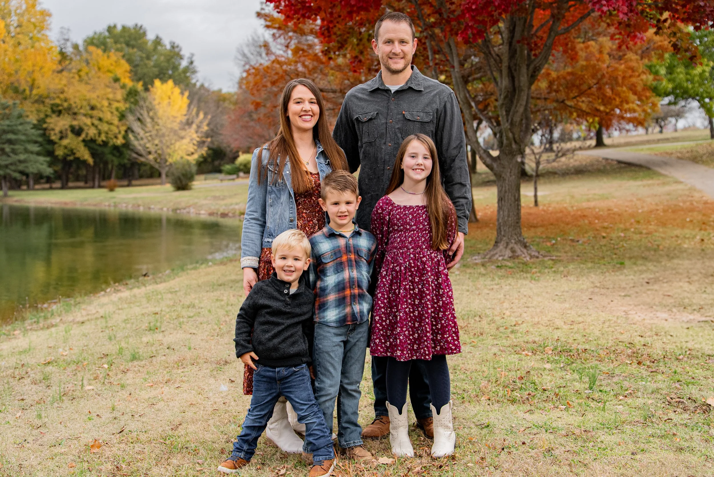 A family of six posed outdoors near a pond with autumn trees in the background. The adults and children are smiling and dressed in casual fall clothing.