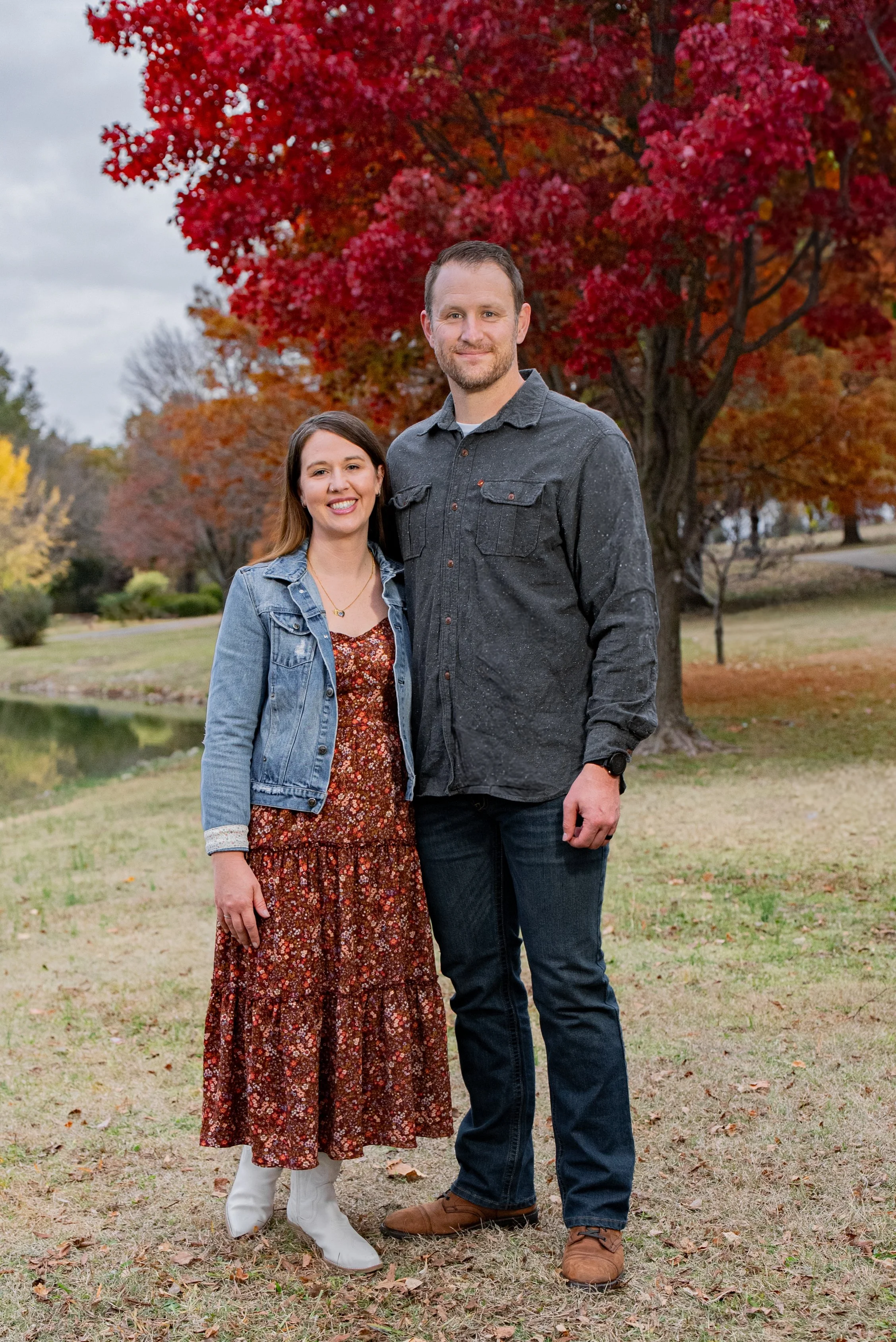 A smiling couple standing outdoors during fall, with colorful red and orange trees in the background, near a small lake.